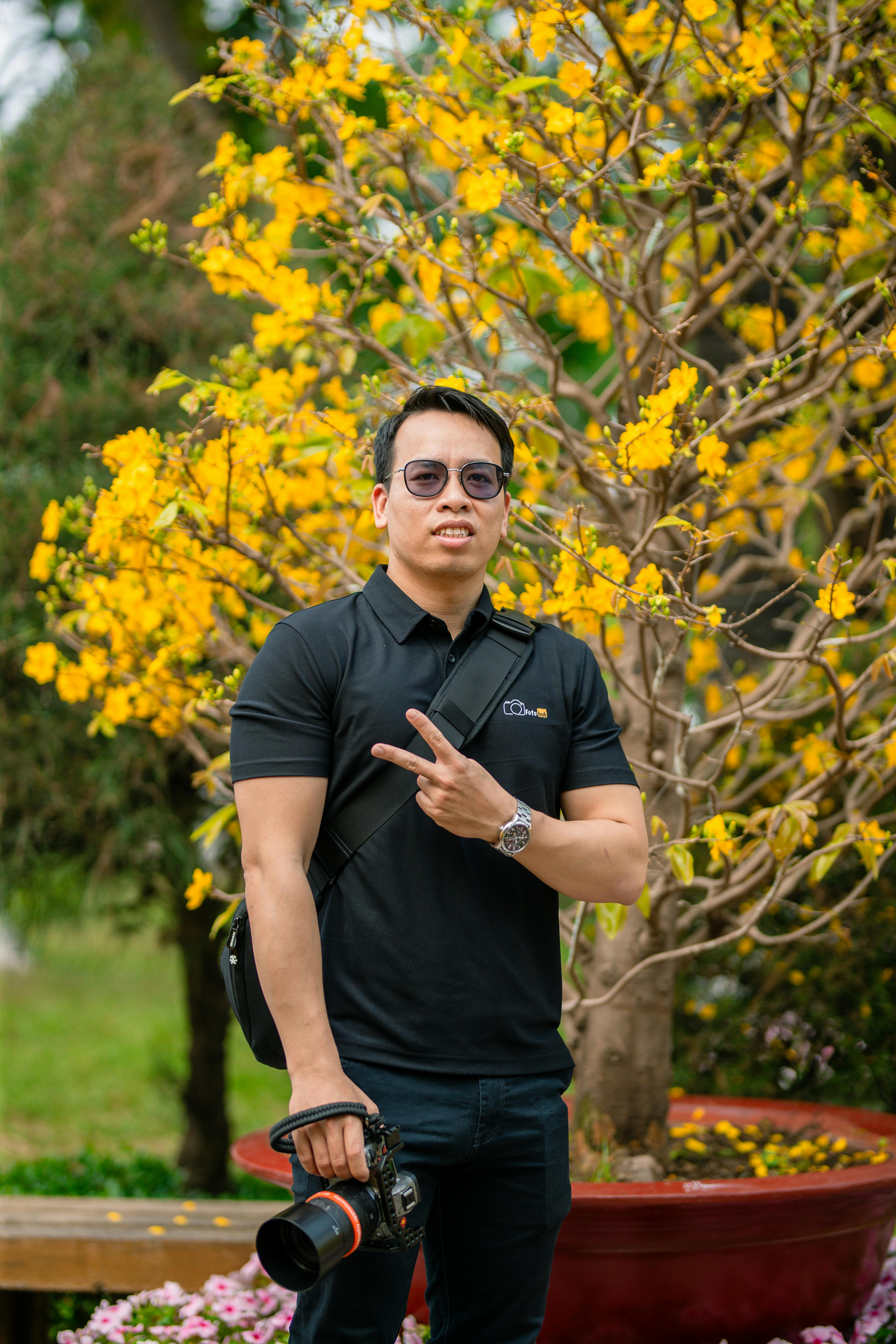 Безкоштовне Чоловік з камерою позує зі знаком миру перед яскравими жовтими квітами в саду. стокове фото