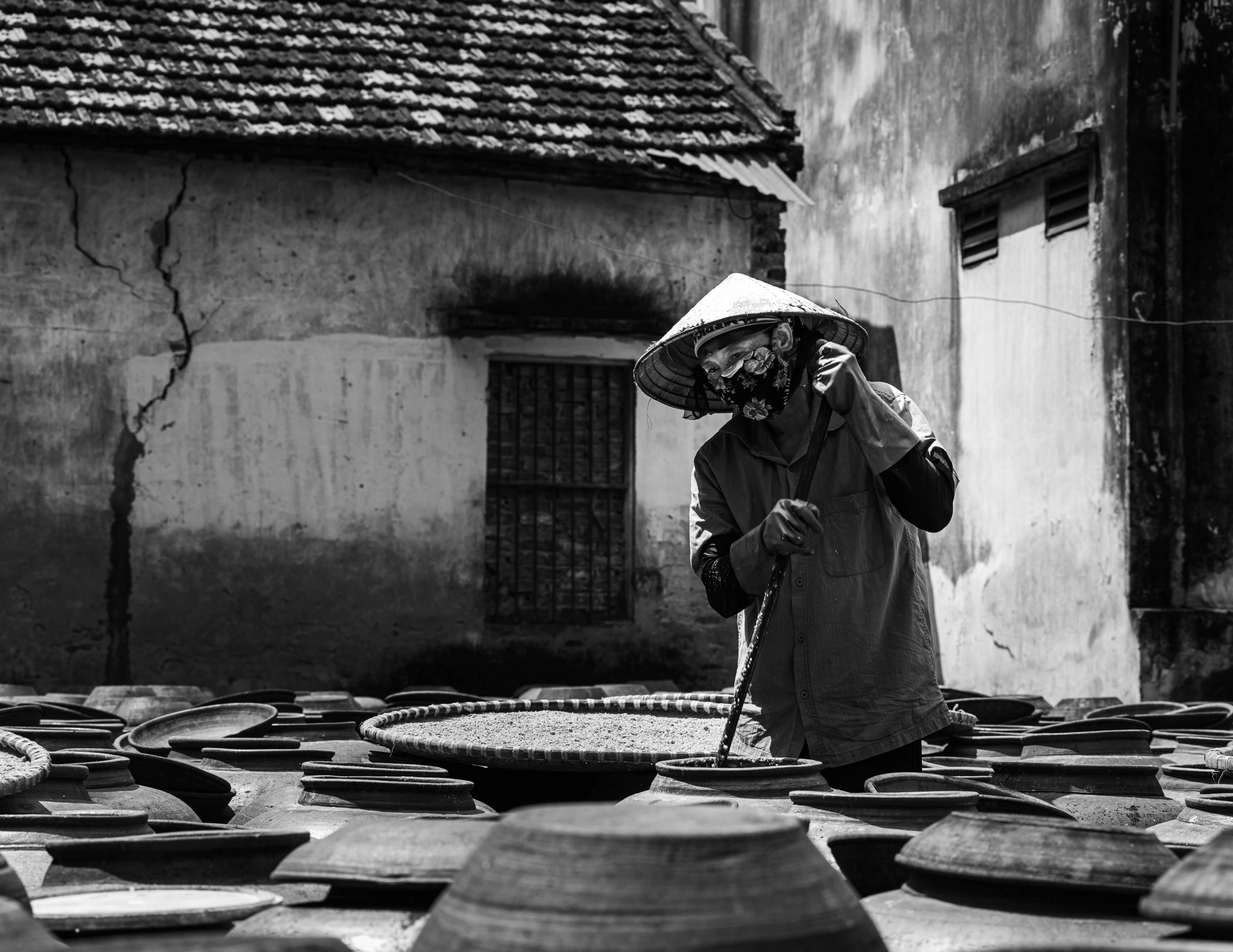 Black and white photo of an artisan in conical hat managing clay pots outdoors.
