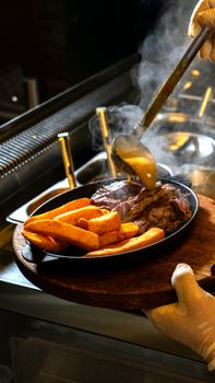 Chef pouring gravy over sizzling steak and fries in kitchen setting.