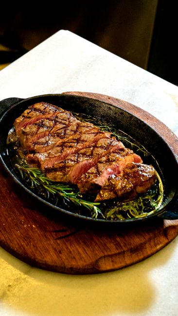 A thick ribeye steak with a dark brown crust resting in a cast iron skillet, being basted with melted butter, garlic cloves, and sprigs of thyme.