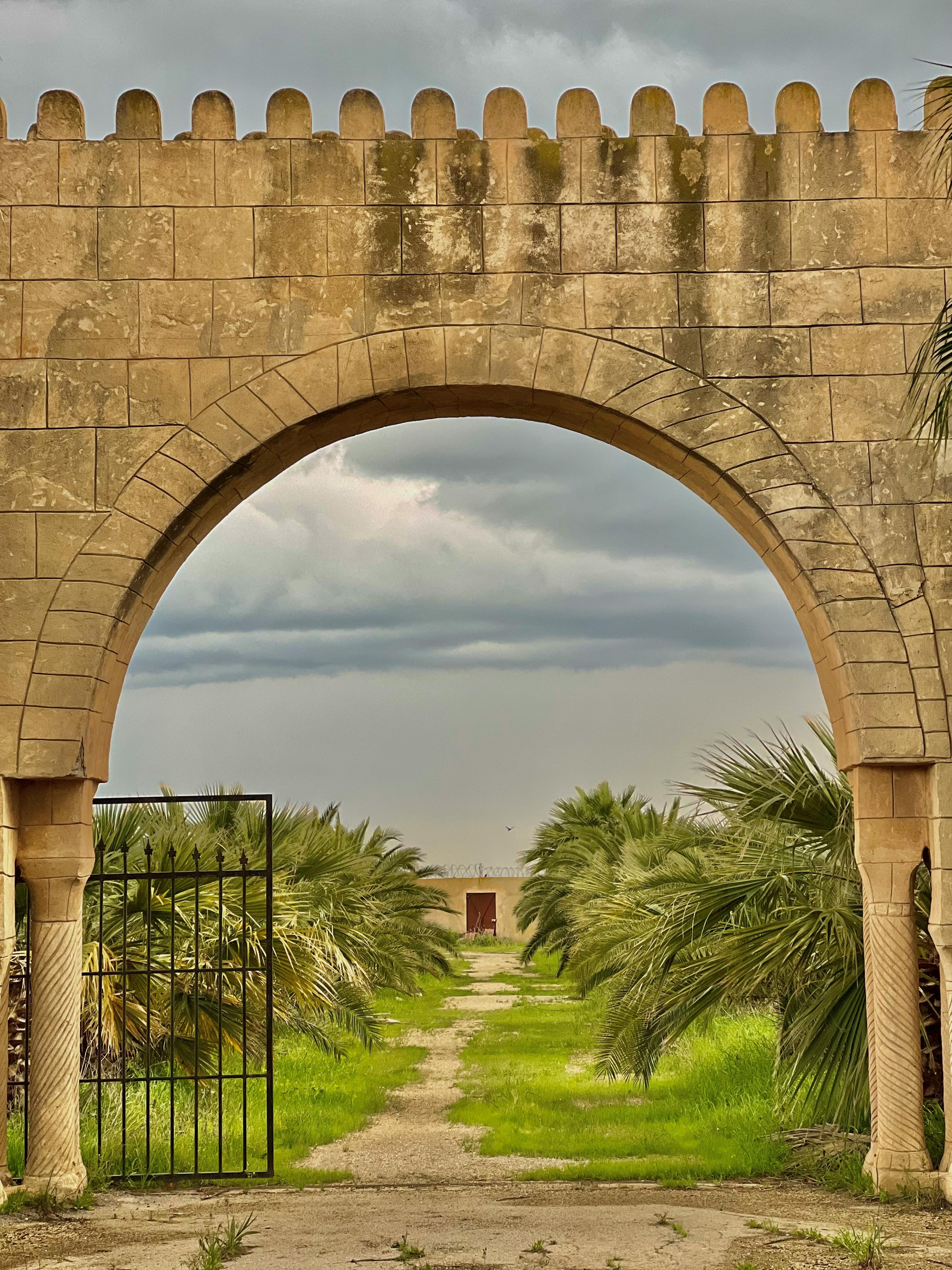 免費 透過古老的石拱門，可以看到棕櫚樹和多雲的天空，景色優美。令人著迷的自然美景。 圖庫相片