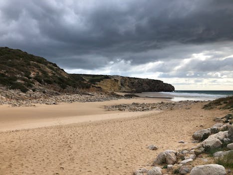 Serene beach scene in Raposeira, Portugal, showcasing sandy shores and a dramatic cloudy sky.