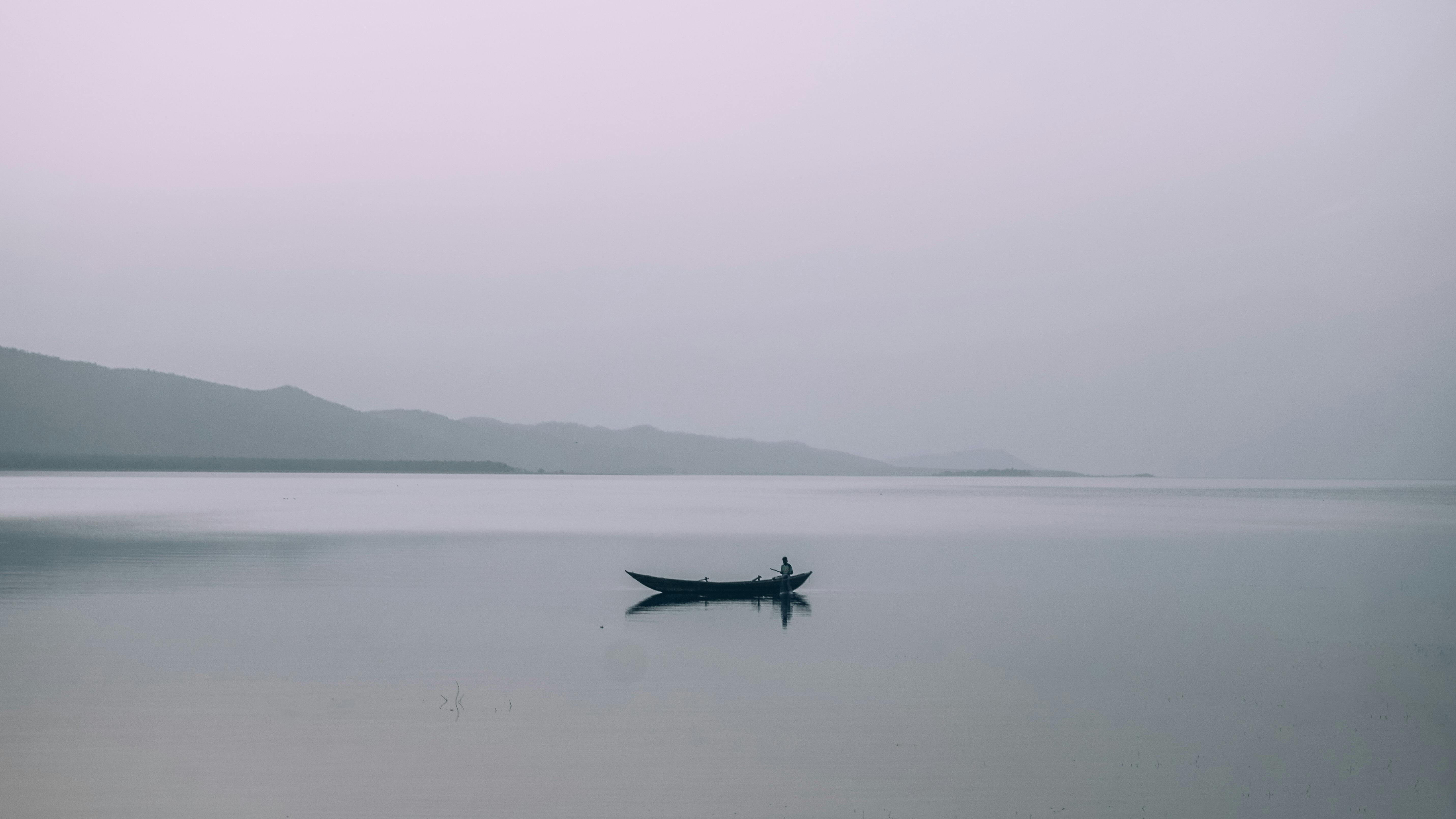 ingyenes Egy magányos csónakázik egy nyugodt tavon, a háttérben ködös hegyekkel alkonyatkor. Stockfotó