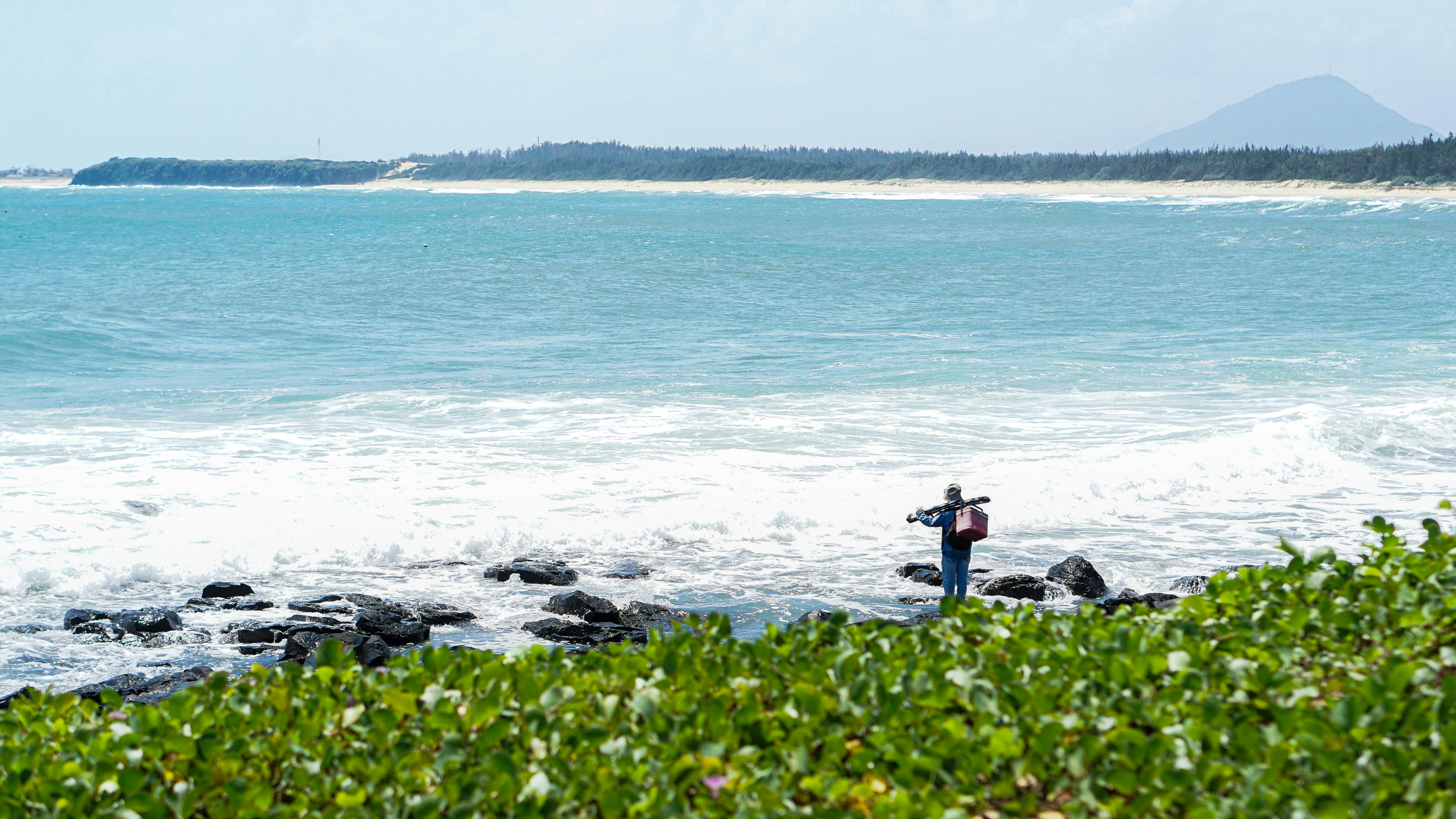 bezplatná Rybář stojí na skalnatém břehu, o který se tříští vlny, pod jasnou oblohou. Základová fotografie
