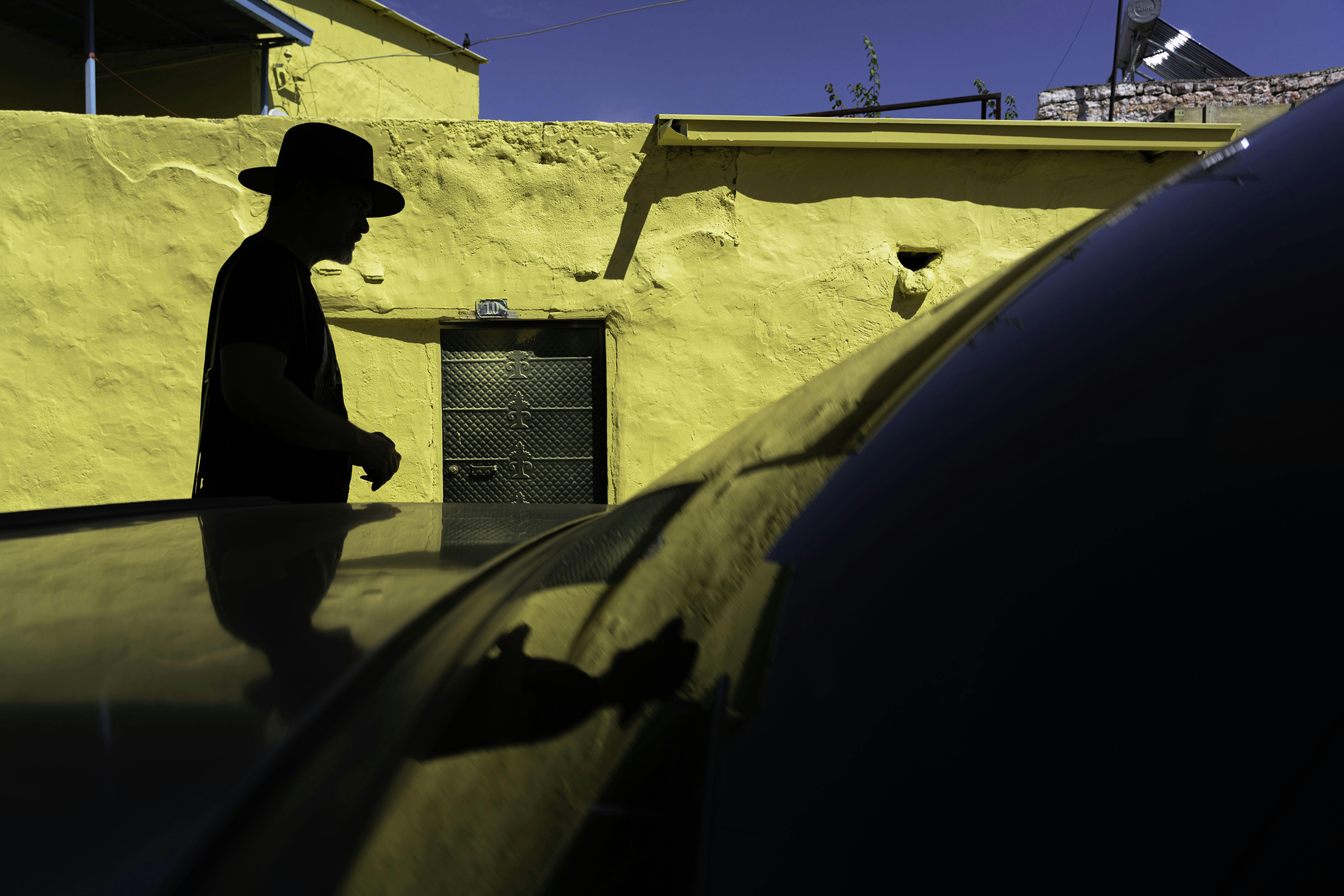 Безкоштовне Силует чоловіка в капелюсі, що стоїть біля жовтої стіни, створюючи контраст з тінями. стокове фото