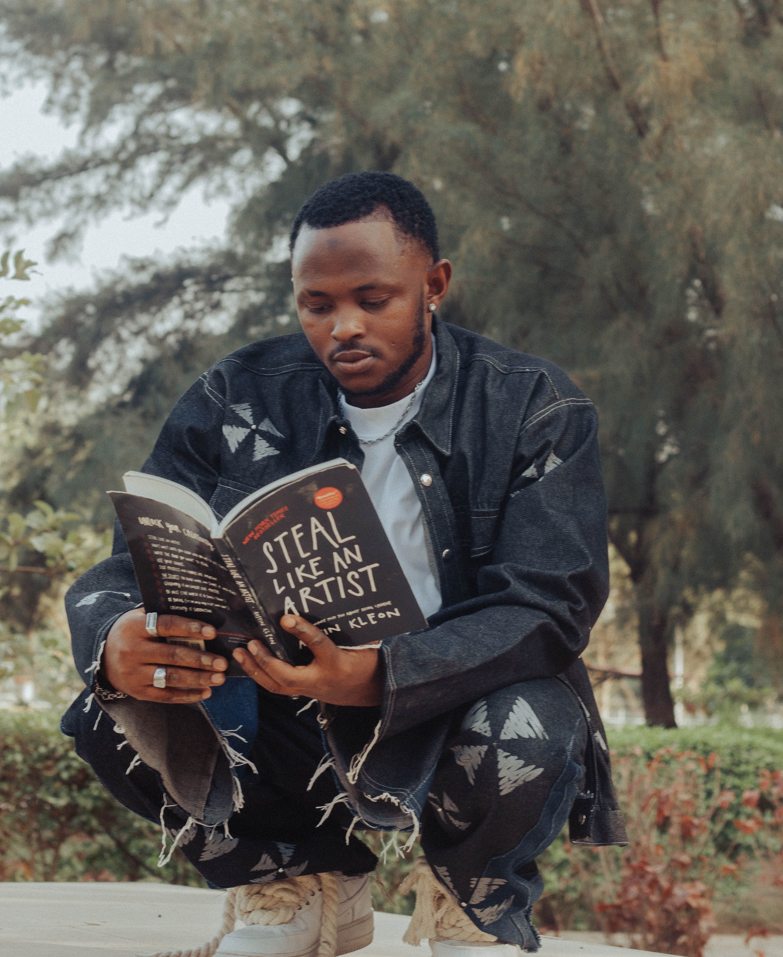 A young man reading 'Steal Like an Artist' outdoors in a park, wearing denim attire.