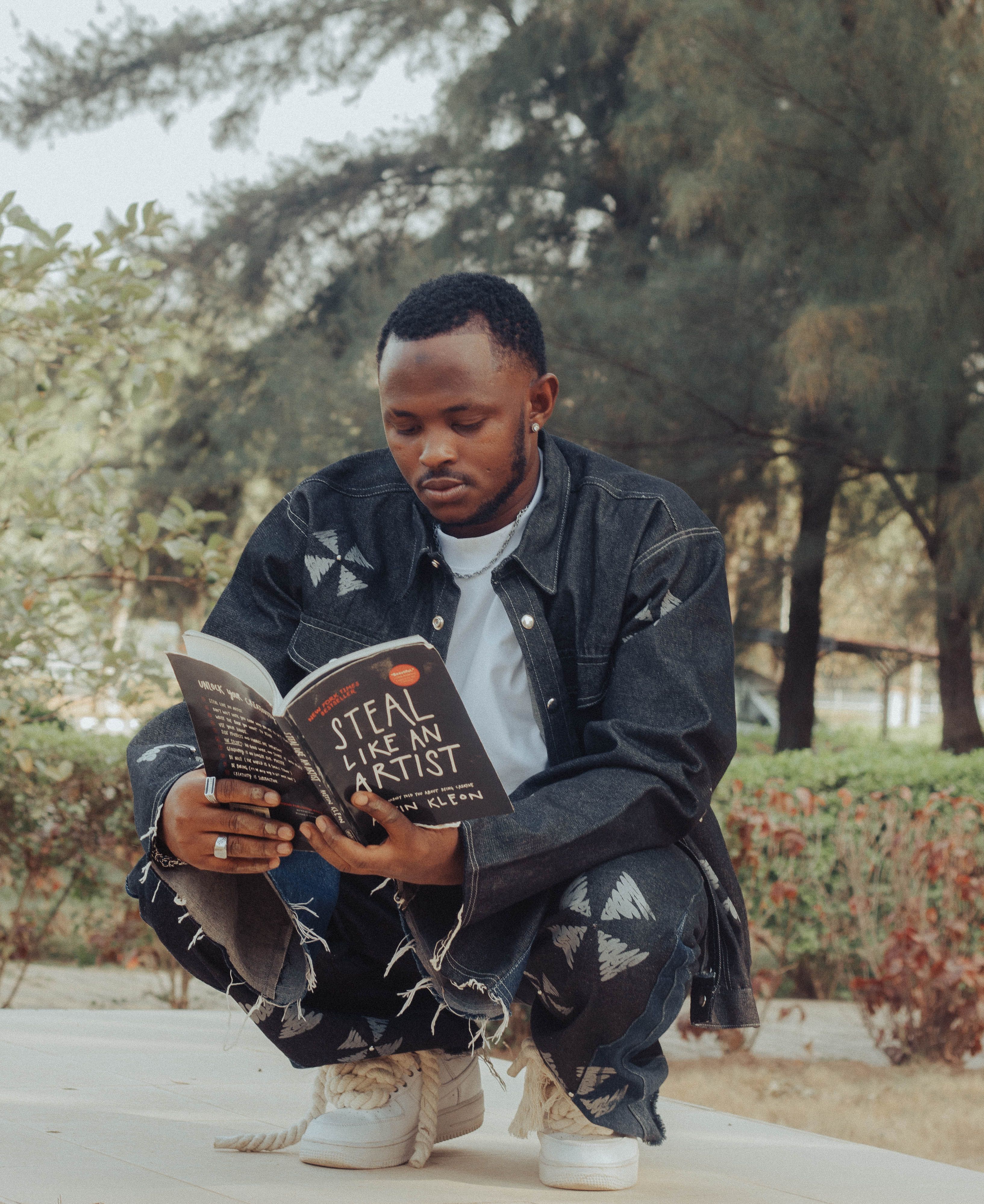 Young man enjoying reading 'Steal Like an Artist' book outdoors in casual denim attire.