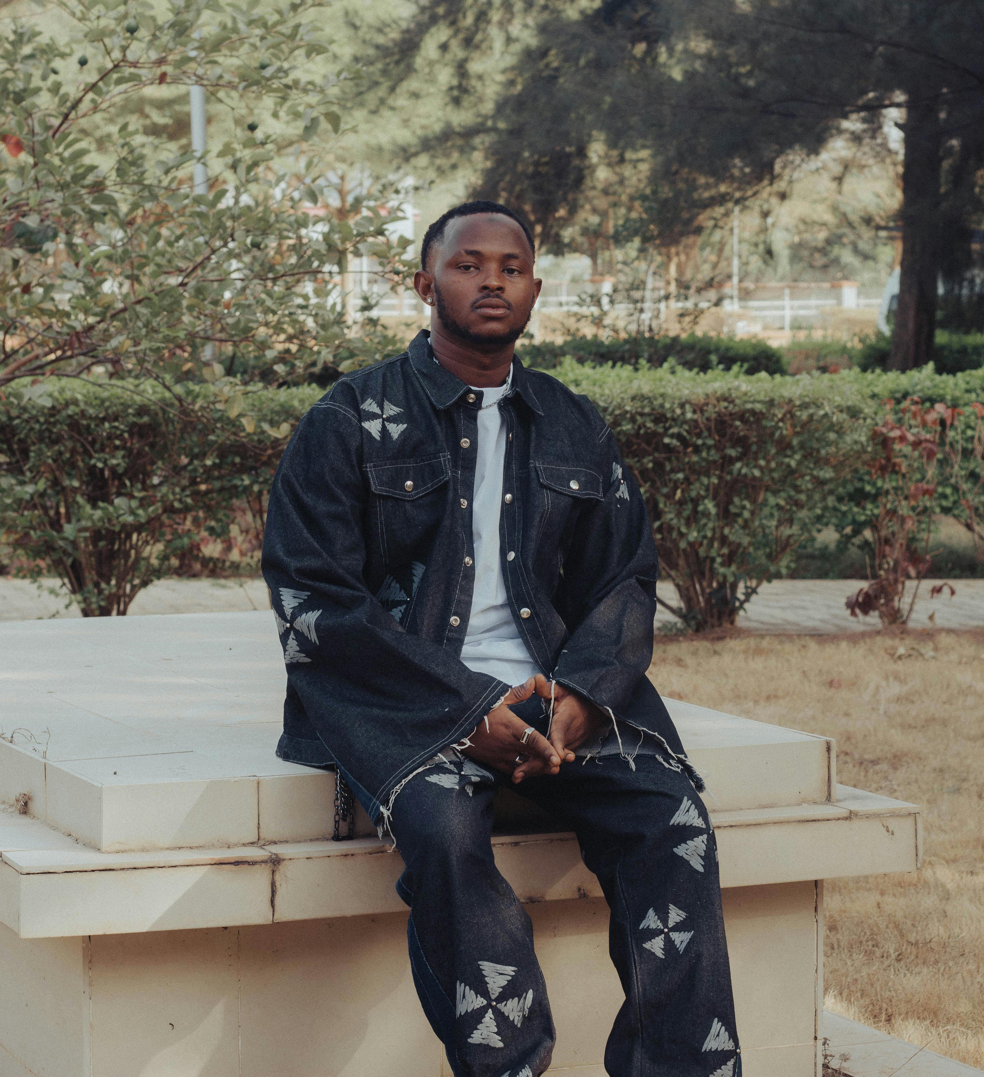 Young man in artistic denim attire poses confidently in a park setting.