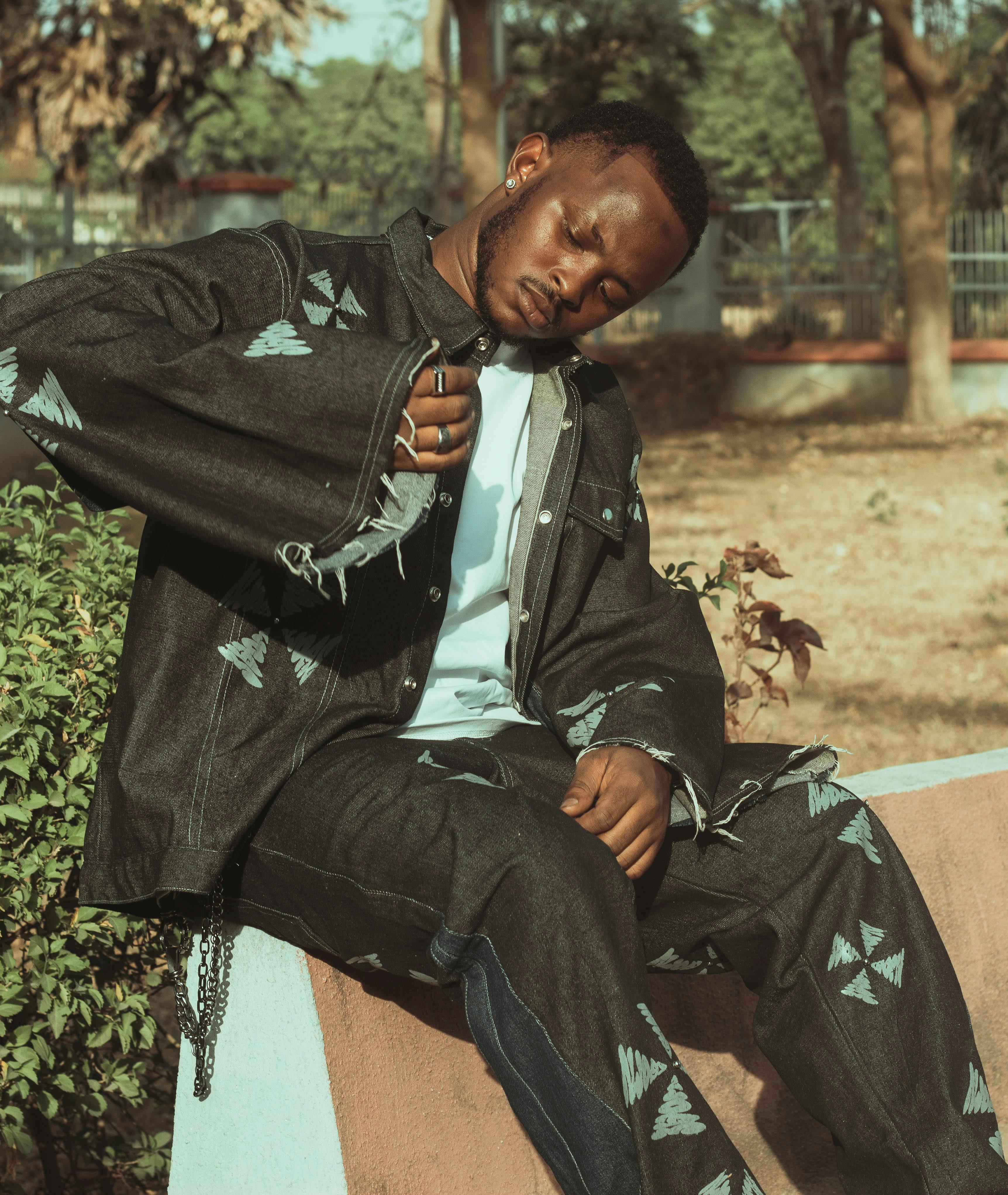 Bezpłatne Młody mężczyzna w wyjątkowym stroju dżinsowym siedzi na zewnątrz, prezentując modny styl. Zdjęcie z galerii