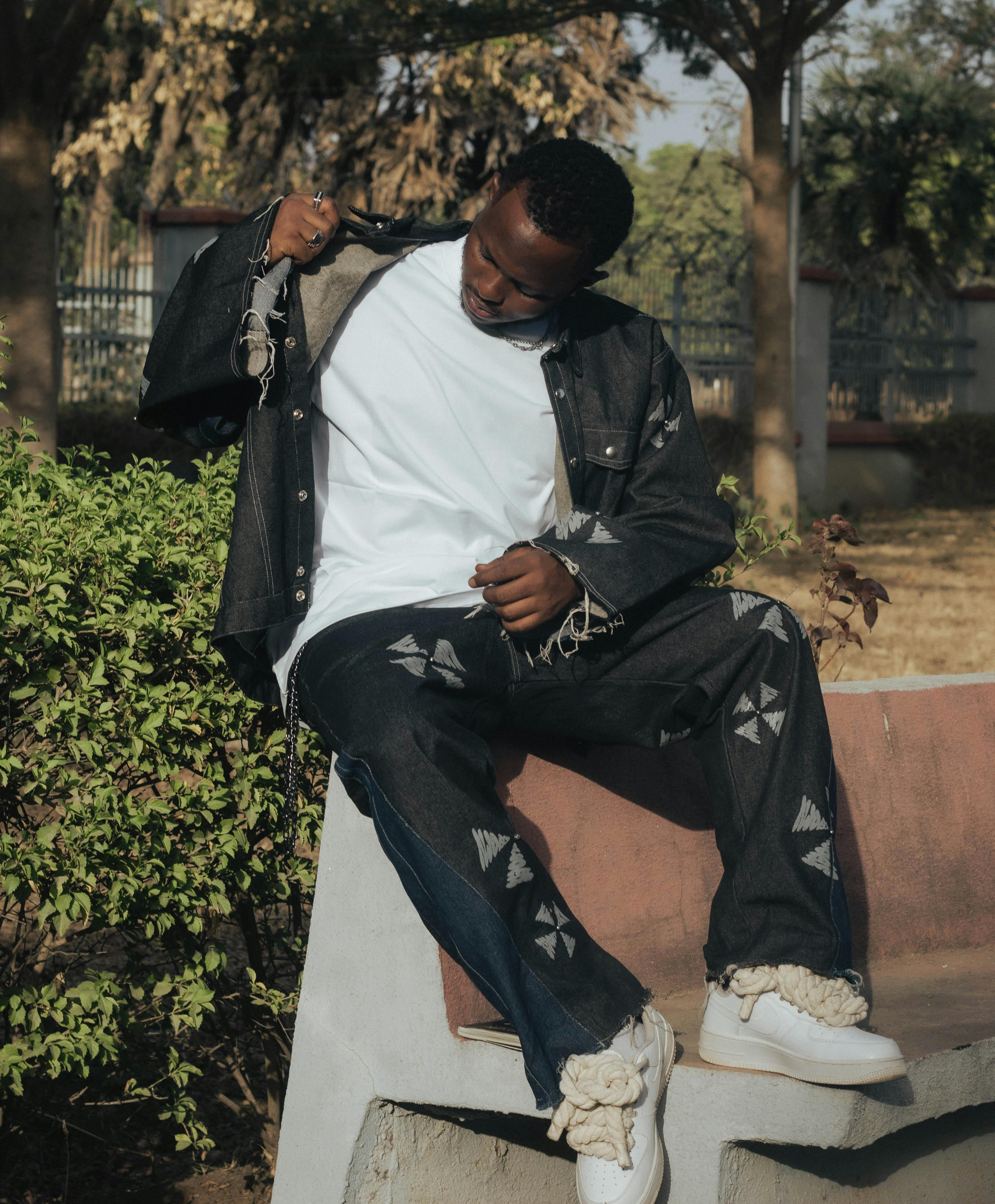 Zadarmo Módny muž v džínsovom oblečení pózuje vonku na lavičke so zeleňou okolo. Fotka z fotobanky