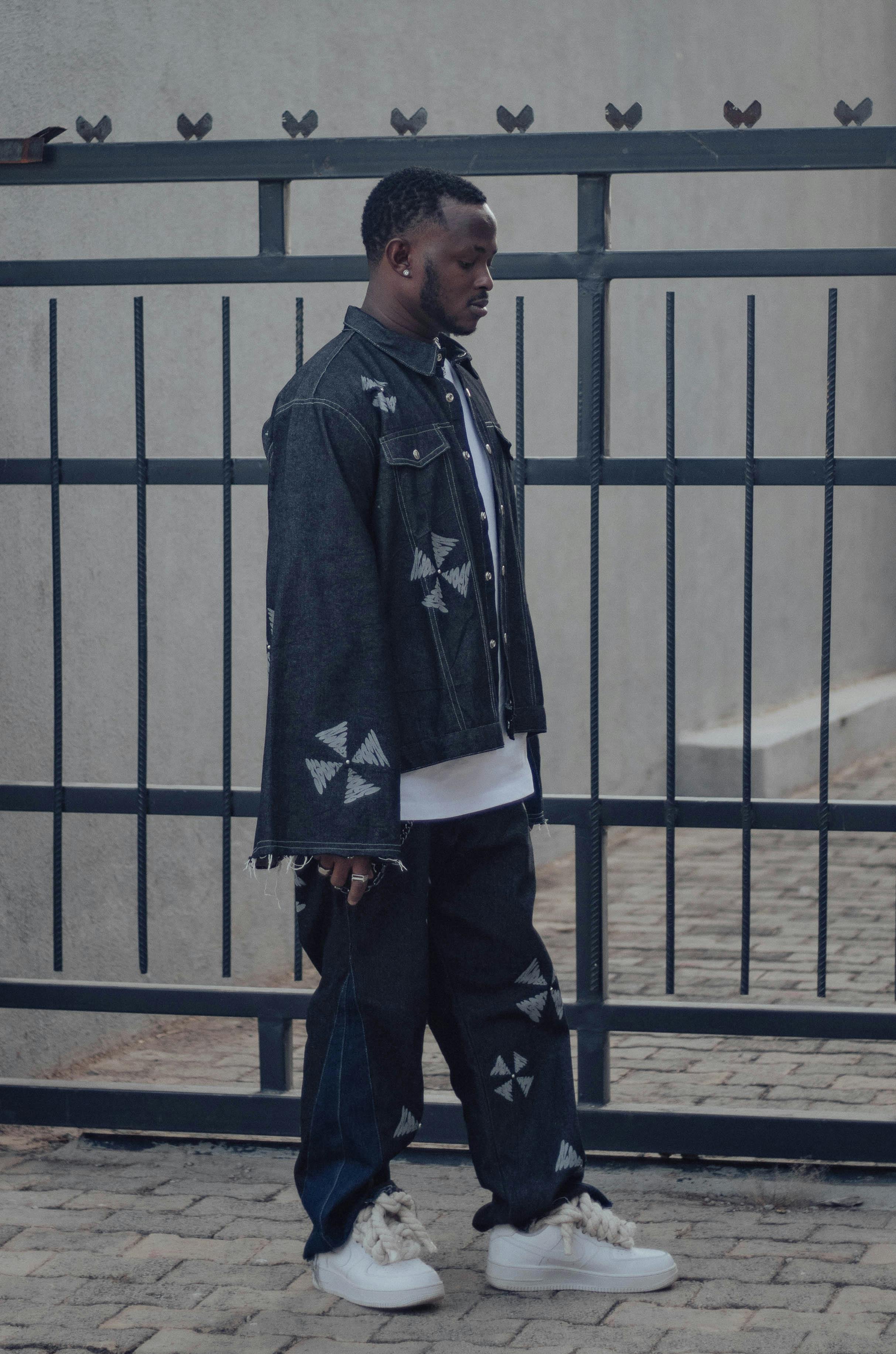 무료 세련된 데님 의상을 입은 젊은 남성이 금속 대문 앞에 서서 도시적인 패션 스타일을 뽐내고 있다. 스톡 사진