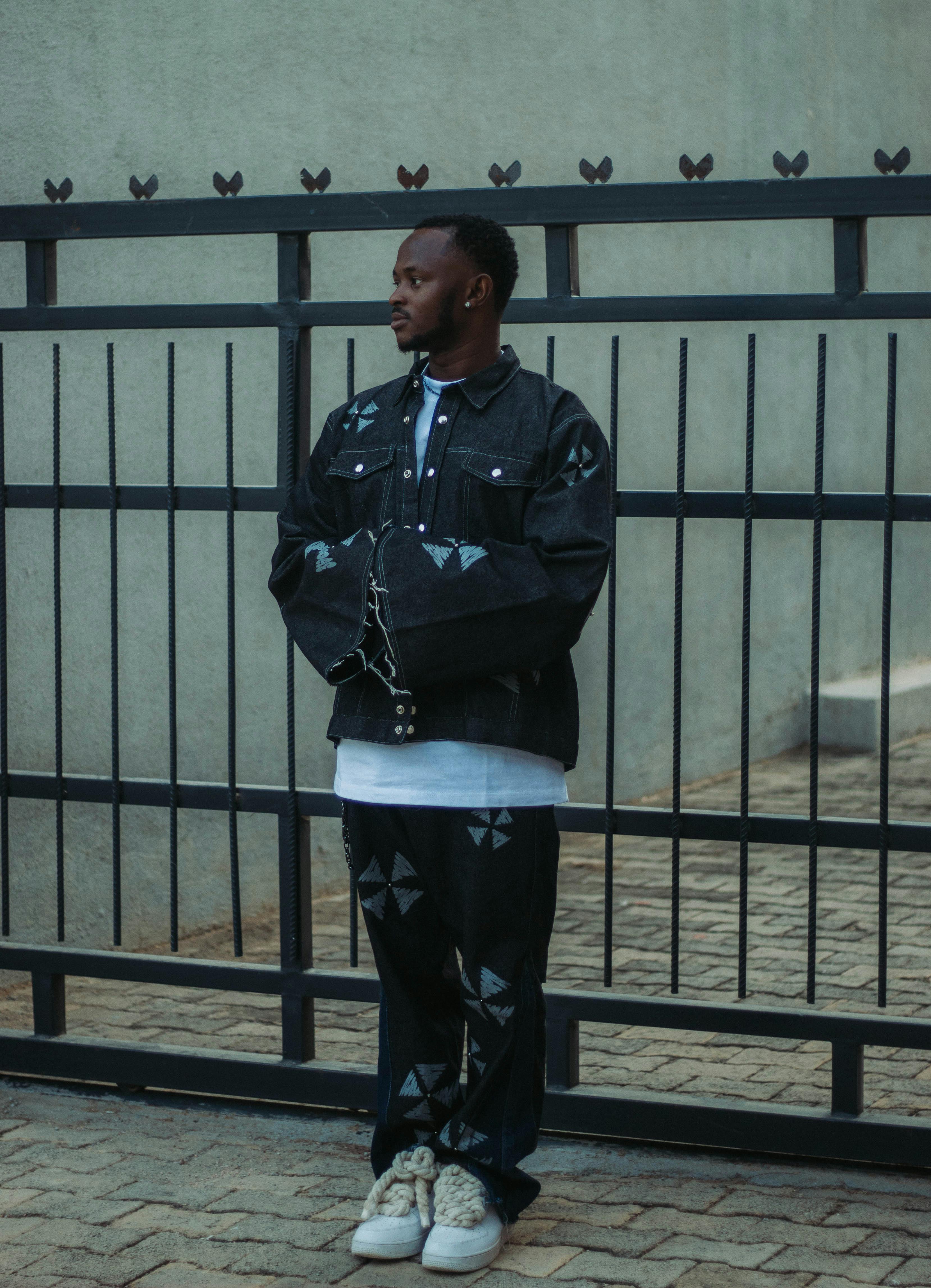 免费 一位身穿深色牛仔夹克和裤子的时尚男士，若有所思地站在城市环境中的一处金属栅栏旁。 素材图片