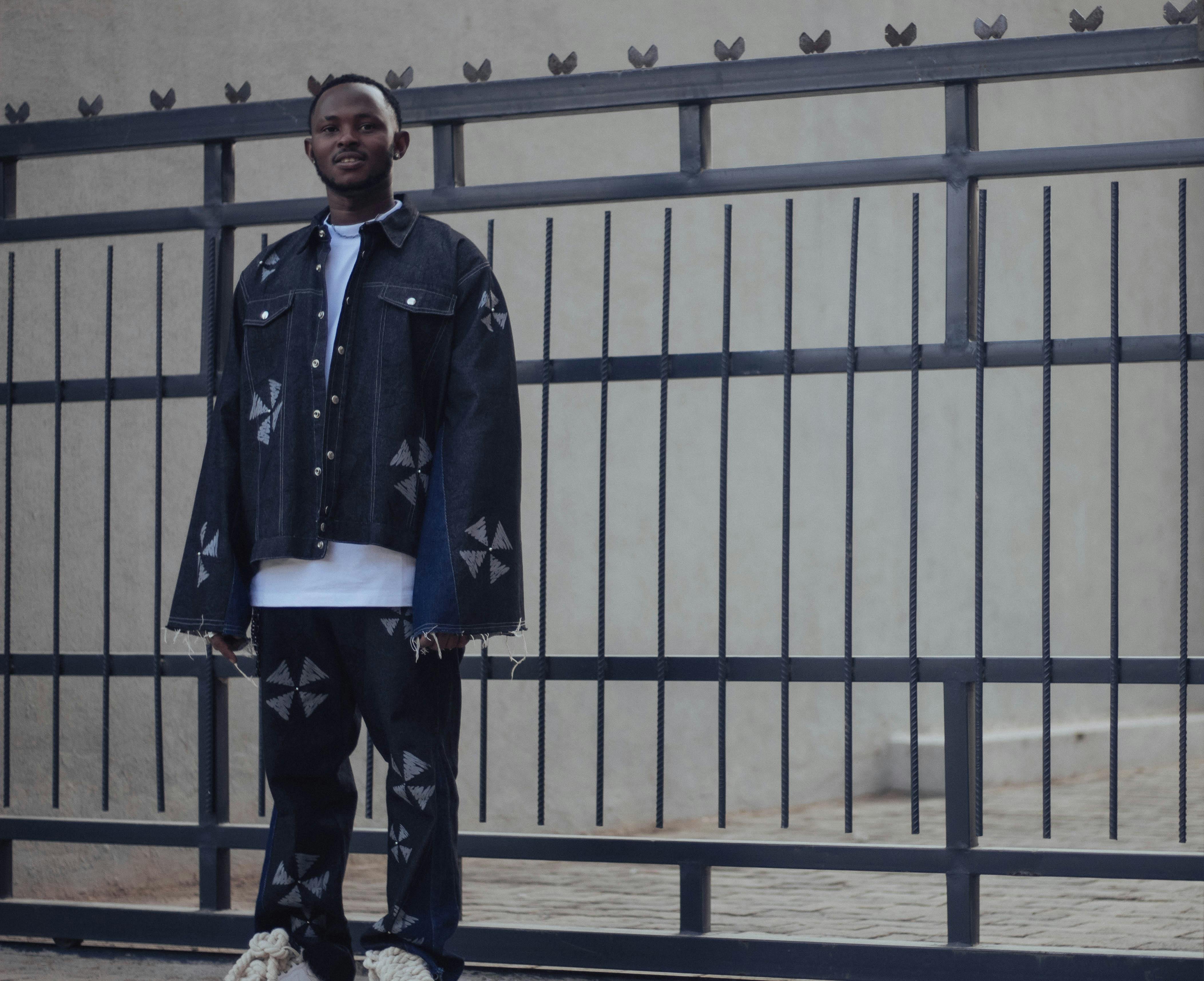 免費 一位身穿時尚牛仔夾克和褲子的年輕男子自信地倚靠在戶外的柵欄上擺姿勢。 圖庫相片