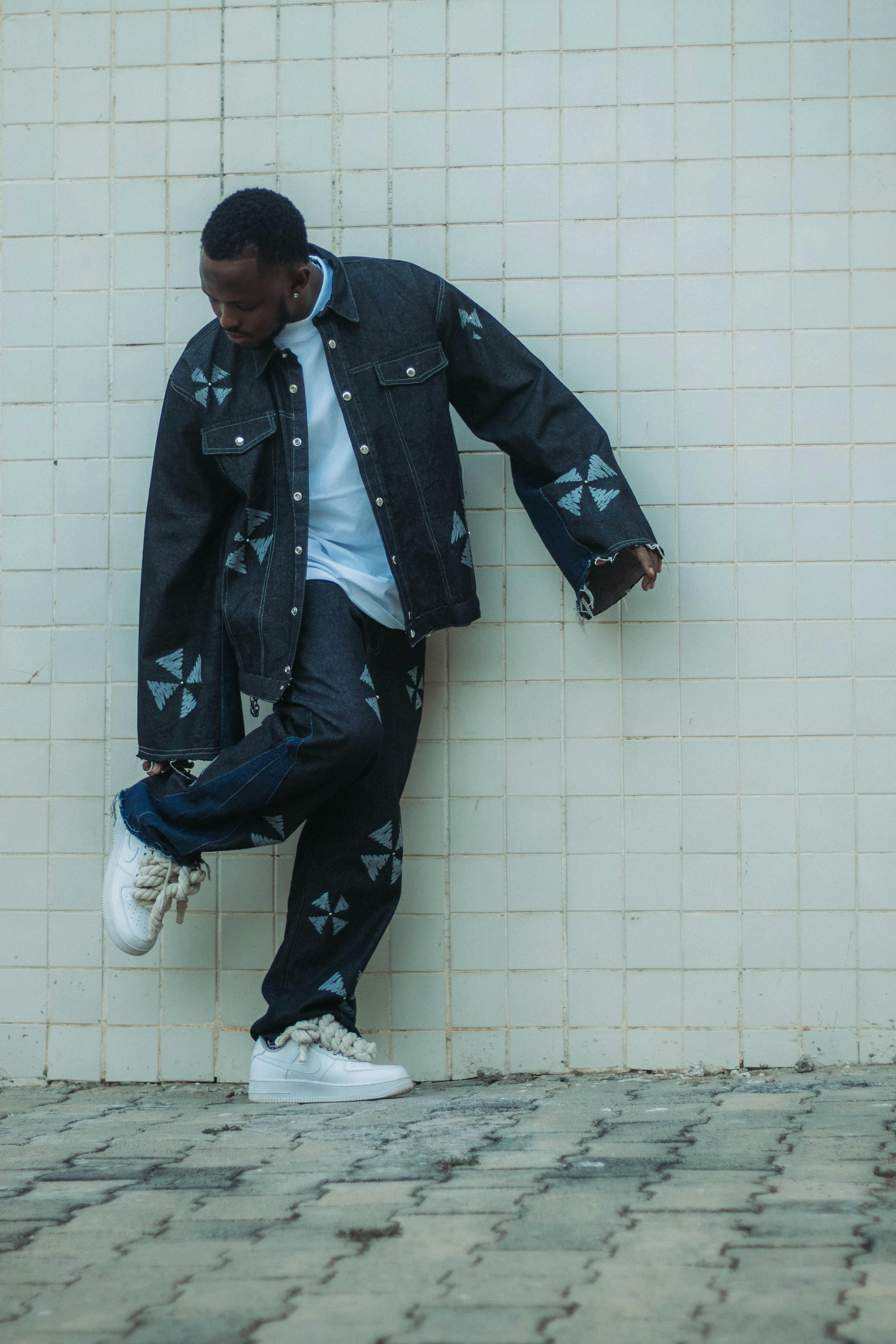 免費 一名年輕男子身穿花式牛仔夾克和褲子，倚靠在瓷磚牆上擺姿勢。 圖庫相片