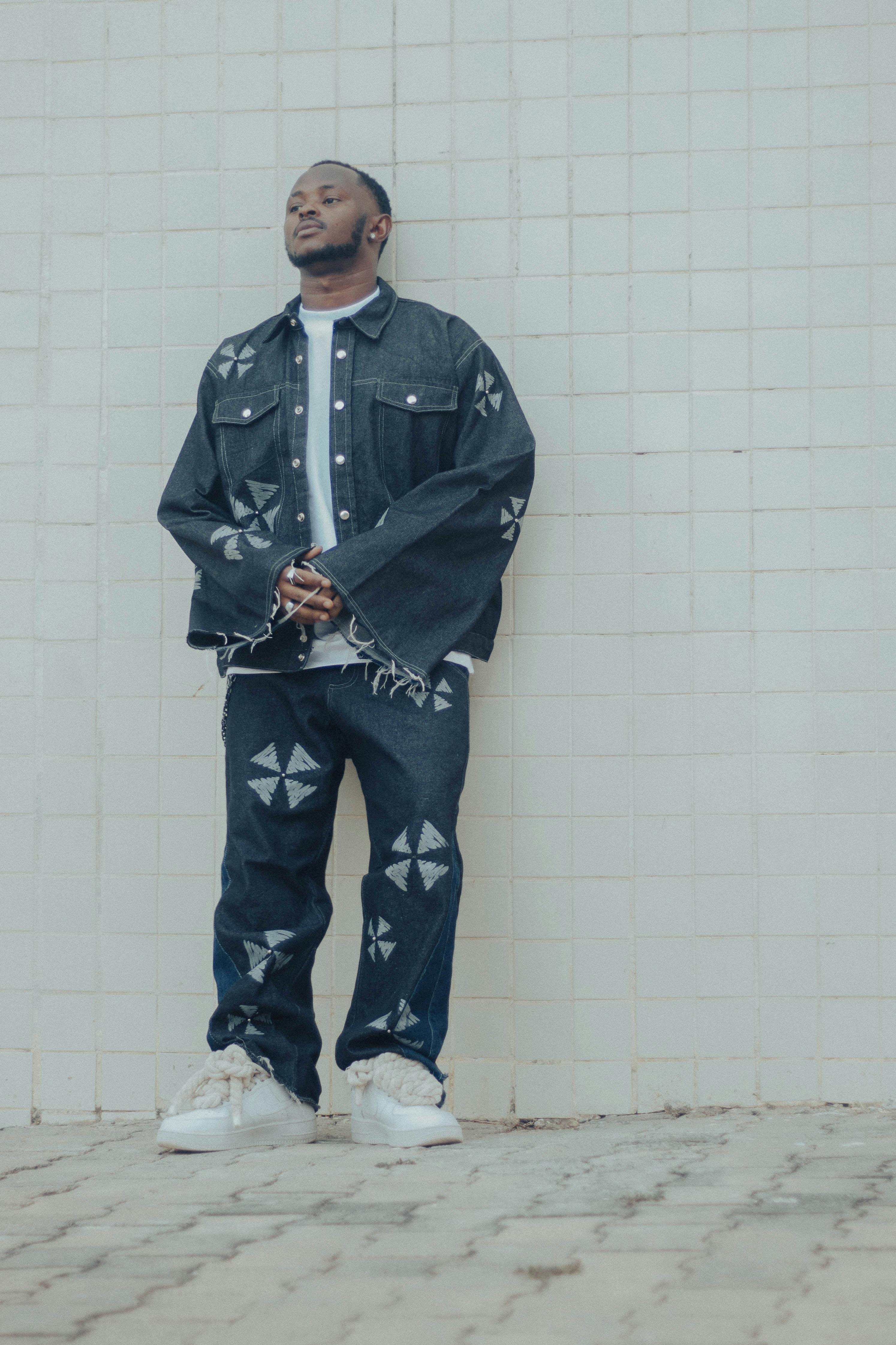 Fashionable young man in denim stands confidently against a textured white wall.