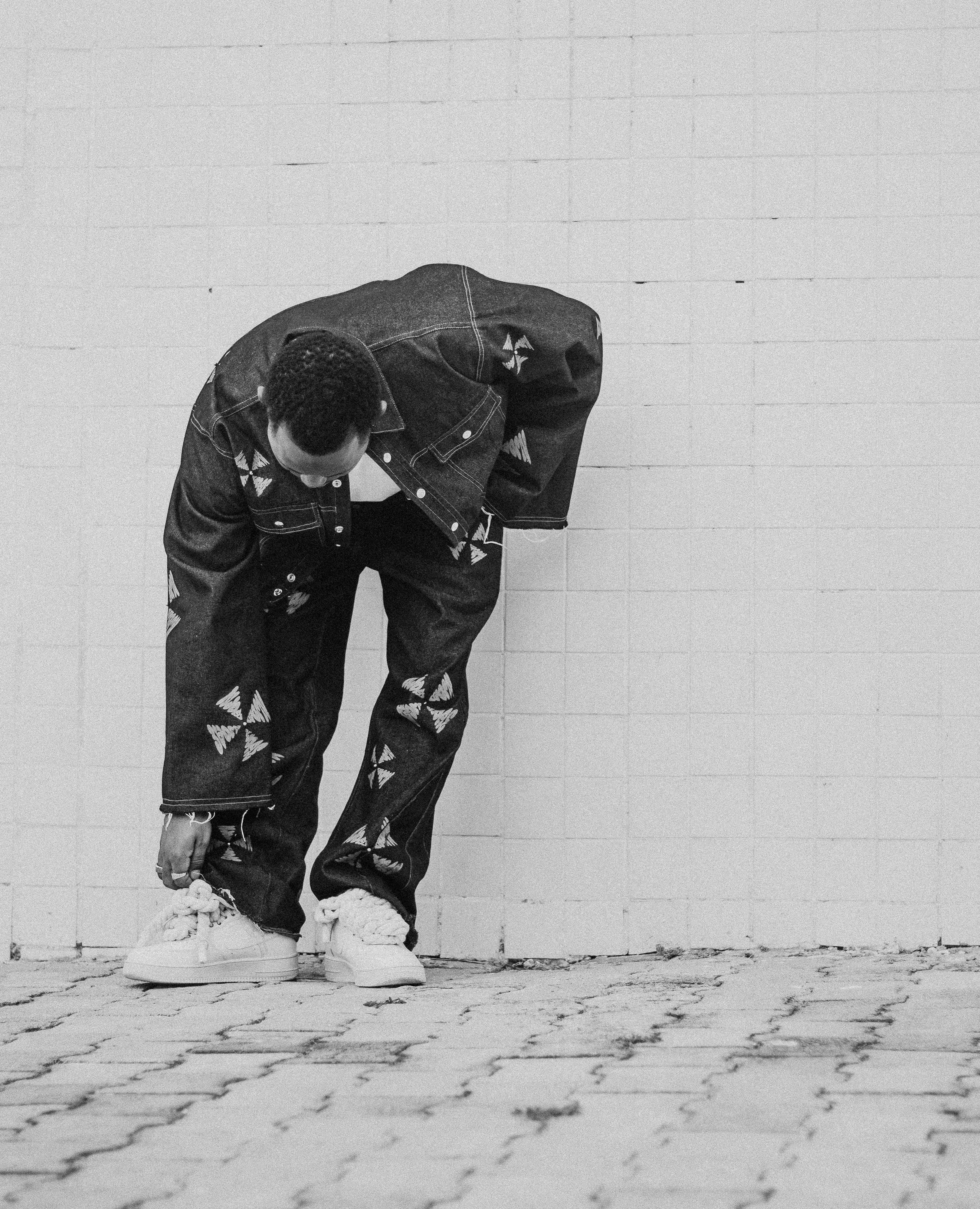 Zadarmo Čiernobiely portrét štýlovej osoby, ktorá si nastavuje šnúrky na topánkach v jedinečnom džínsovom oblečení pri obloženej stene. Fotka z fotobanky