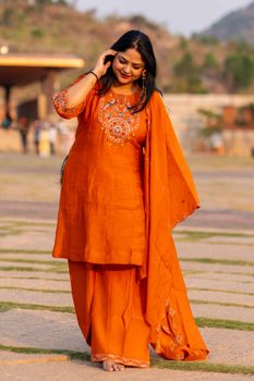Smiling woman in orange ethnic attire walking outdoors with mountains in the background.