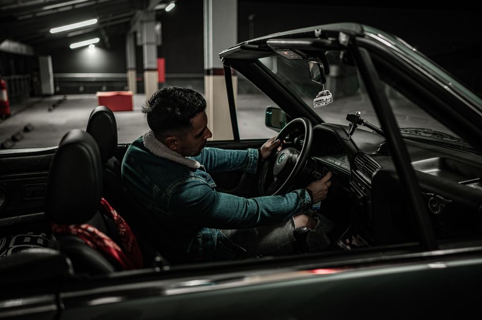 A stylish adult male driving a convertible car in a dimly lit underground parking garage.
