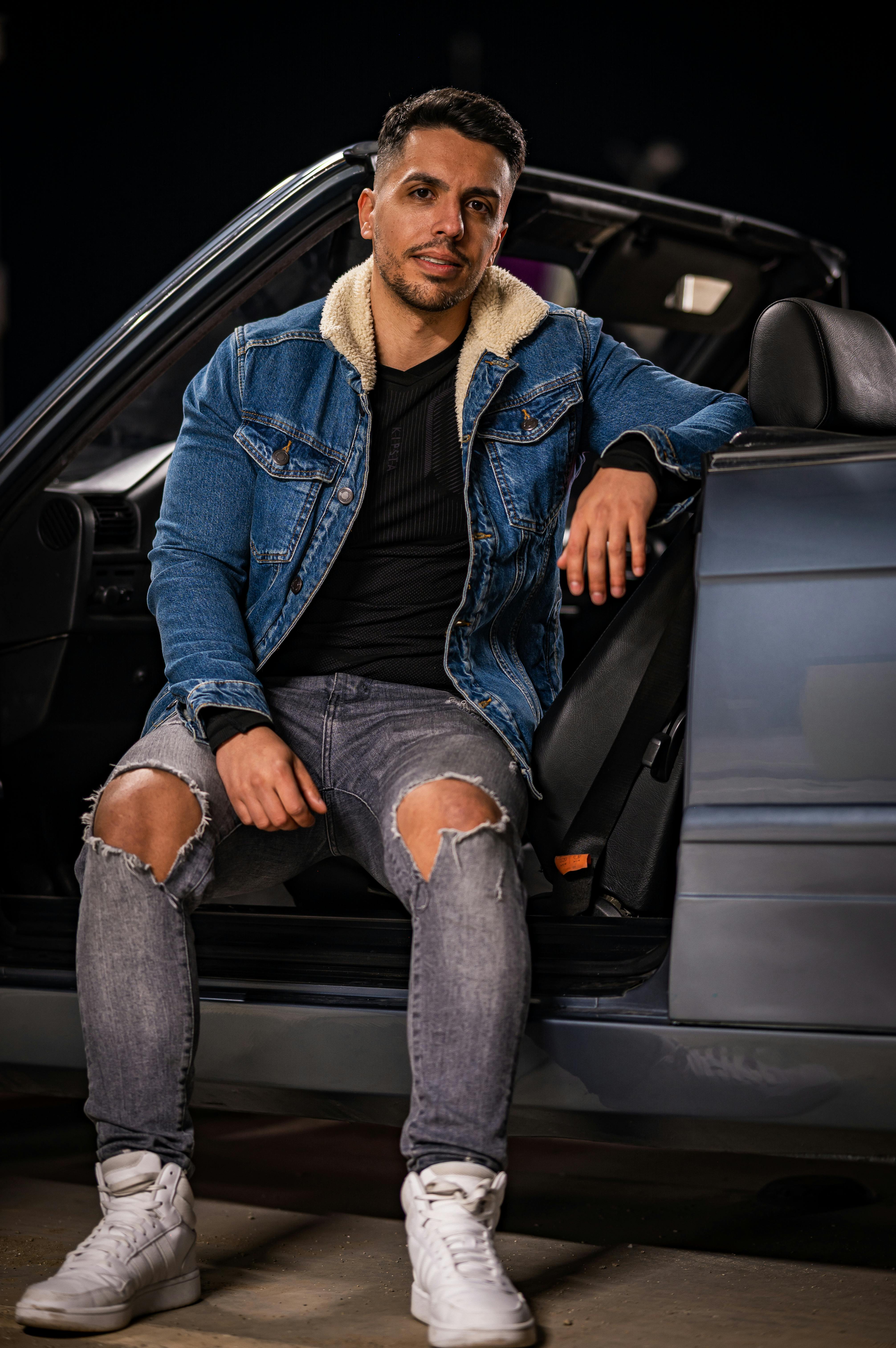 免費 夜晚，一位身穿牛仔褲和運動鞋的年輕男子靠在一輛敞篷車上。 圖庫相片