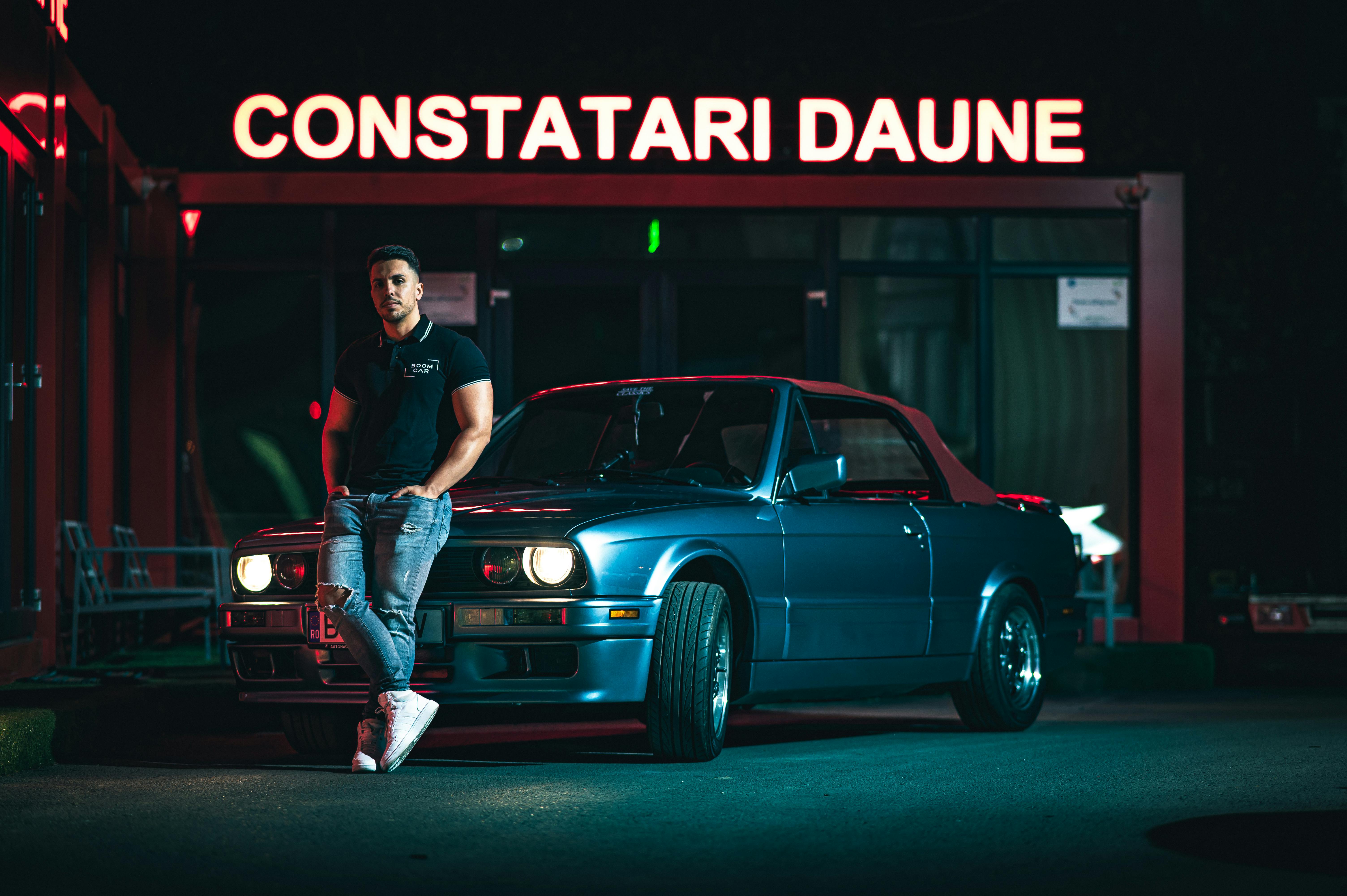 ฟรี ชายหนุ่มผู้มีสไตล์ยืนพิงรถยนต์สีฟ้าโบราณใต้แสงไฟนีออนในเวลากลางคืน คลังภาพถ่าย