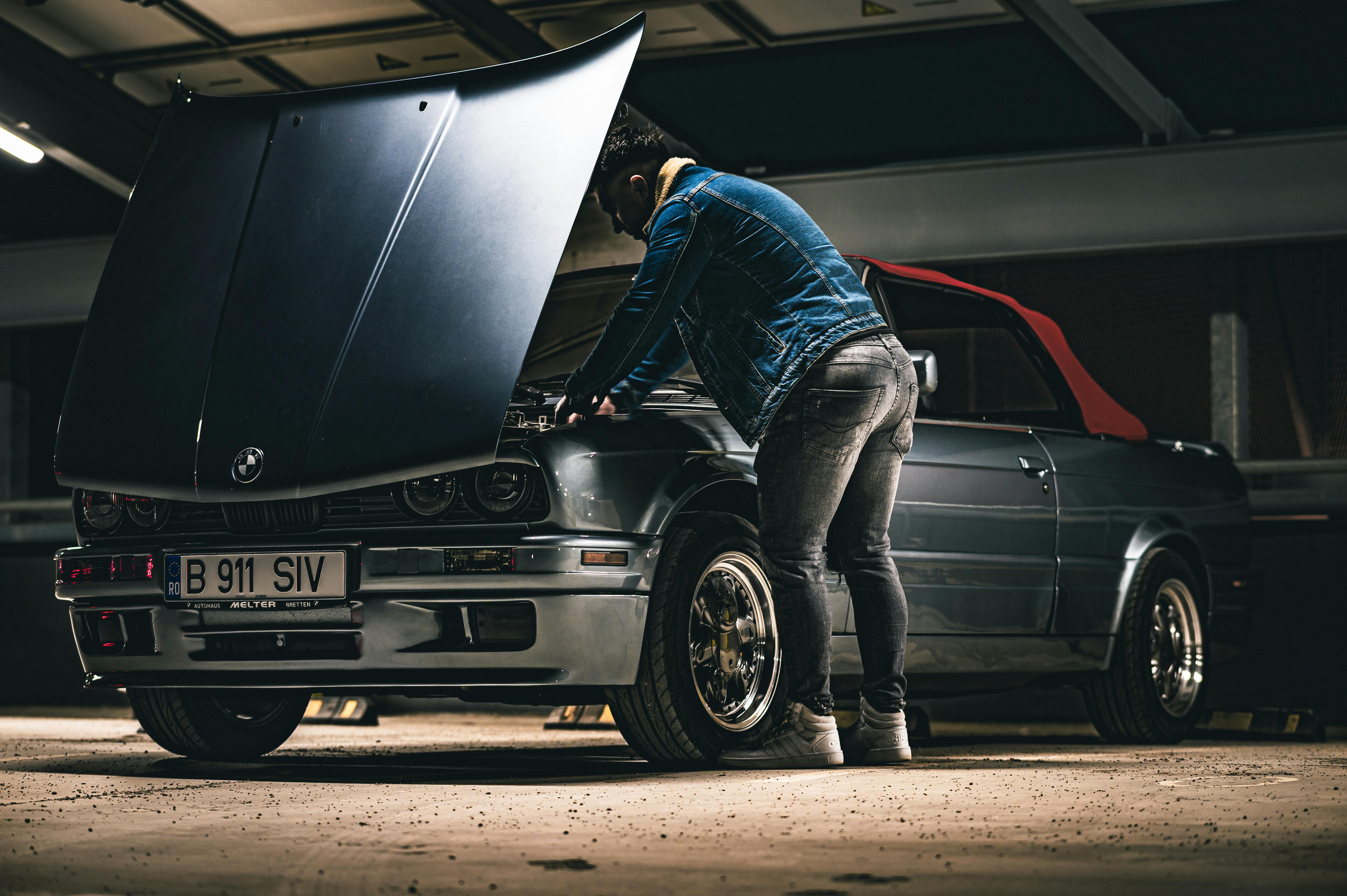 무료 데님 재킷을 입은 남자가 어둑한 차고 안에서 클래식 컨버터블 자동차를 수리하고 있다. 스톡 사진