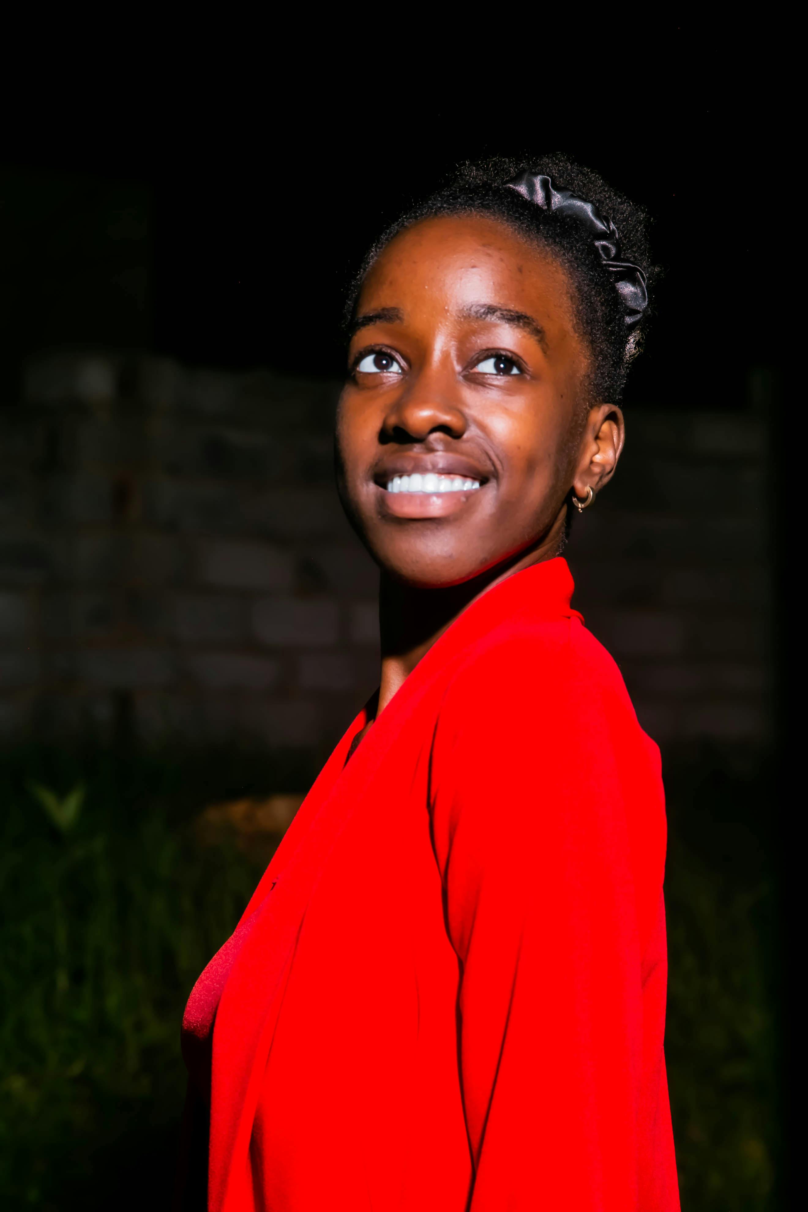 ฟรี หญิงสาวคนหนึ่งยิ้มอย่างมั่นใจในเวลากลางคืน สวมเสื้อแจ็กเก็ตสีแดงสดตัดกับฉากหลังสีเข้ม คลังภาพถ่าย