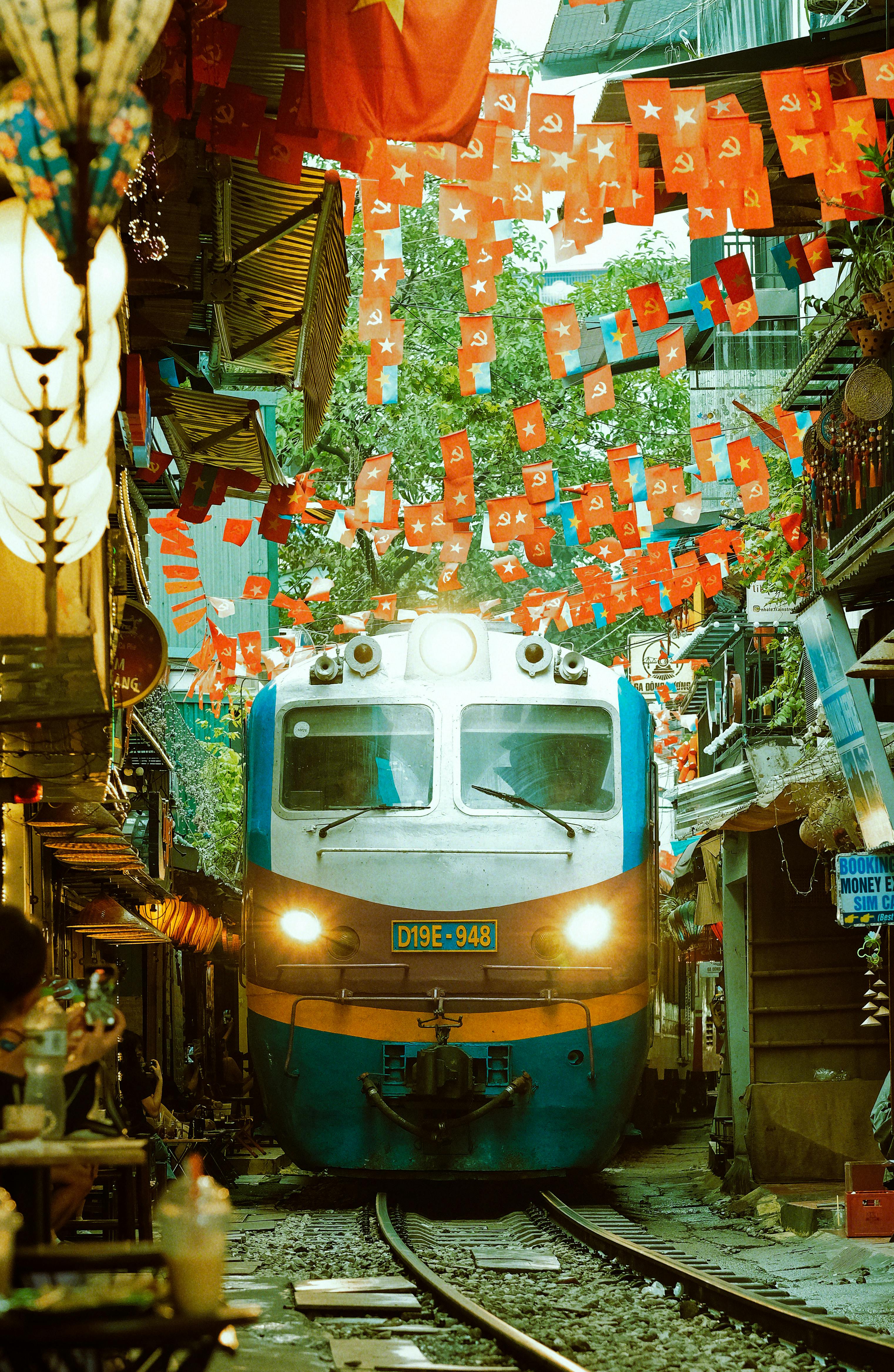 ฟรี รถไฟหลากสีสันแล่นผ่านตลาดริมถนนที่คึกคักของกรุงฮานอย ประเทศเวียดนาม ซึ่งประดับประดาด้วยธงสีสันสดใส คลังภาพถ่าย