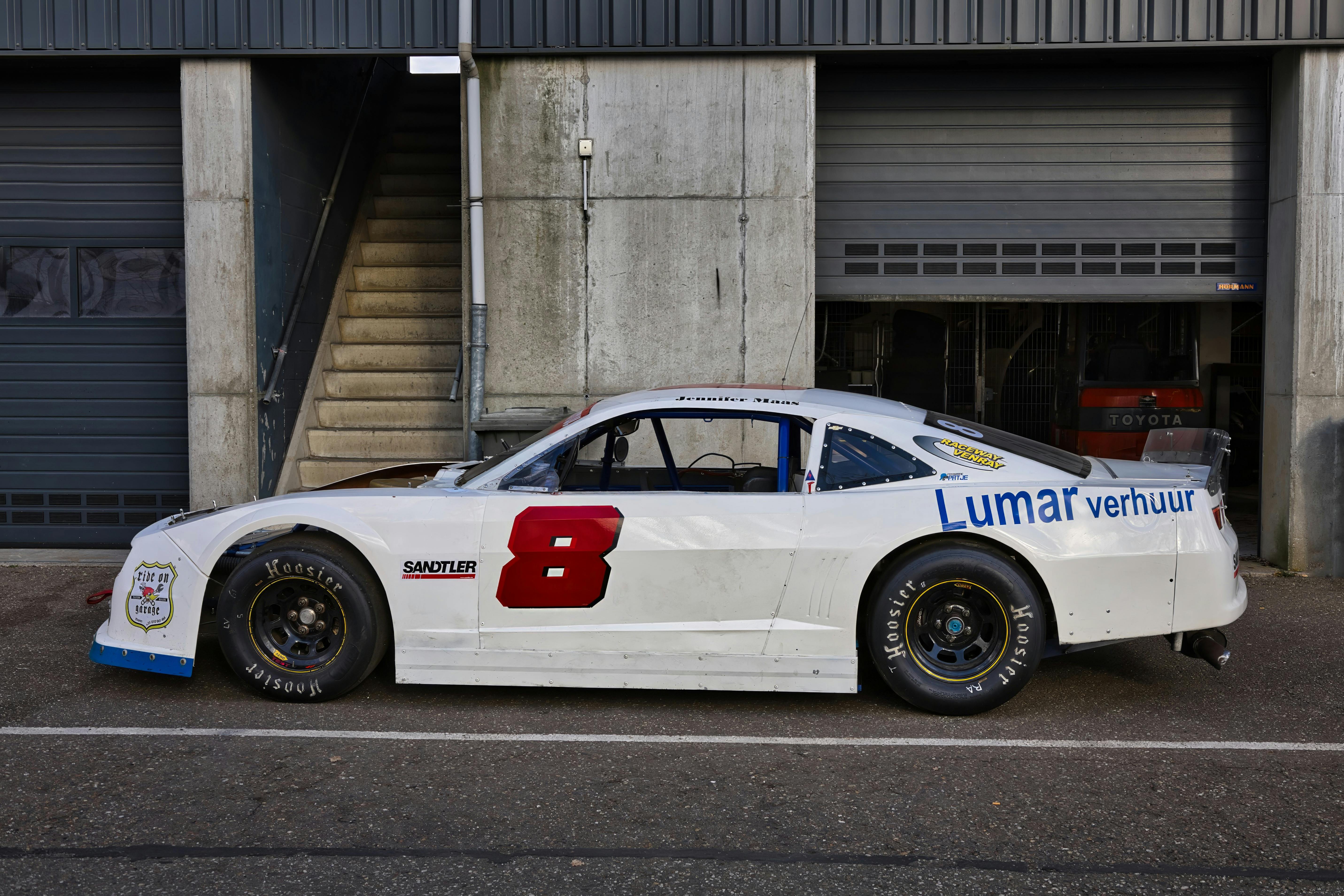 ฟรี รถแข่งสีขาวจอดอยู่กลางแจ้งใกล้อู่ซ่อมรถในวันที่แดดจ้า คลังภาพถ่าย