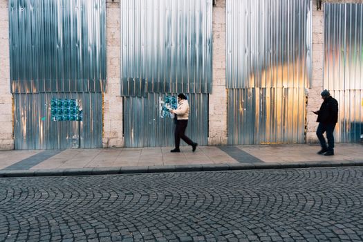 Two pedestrians walk along a metal-paneled street in Istanbul, Türkiye, on a day.