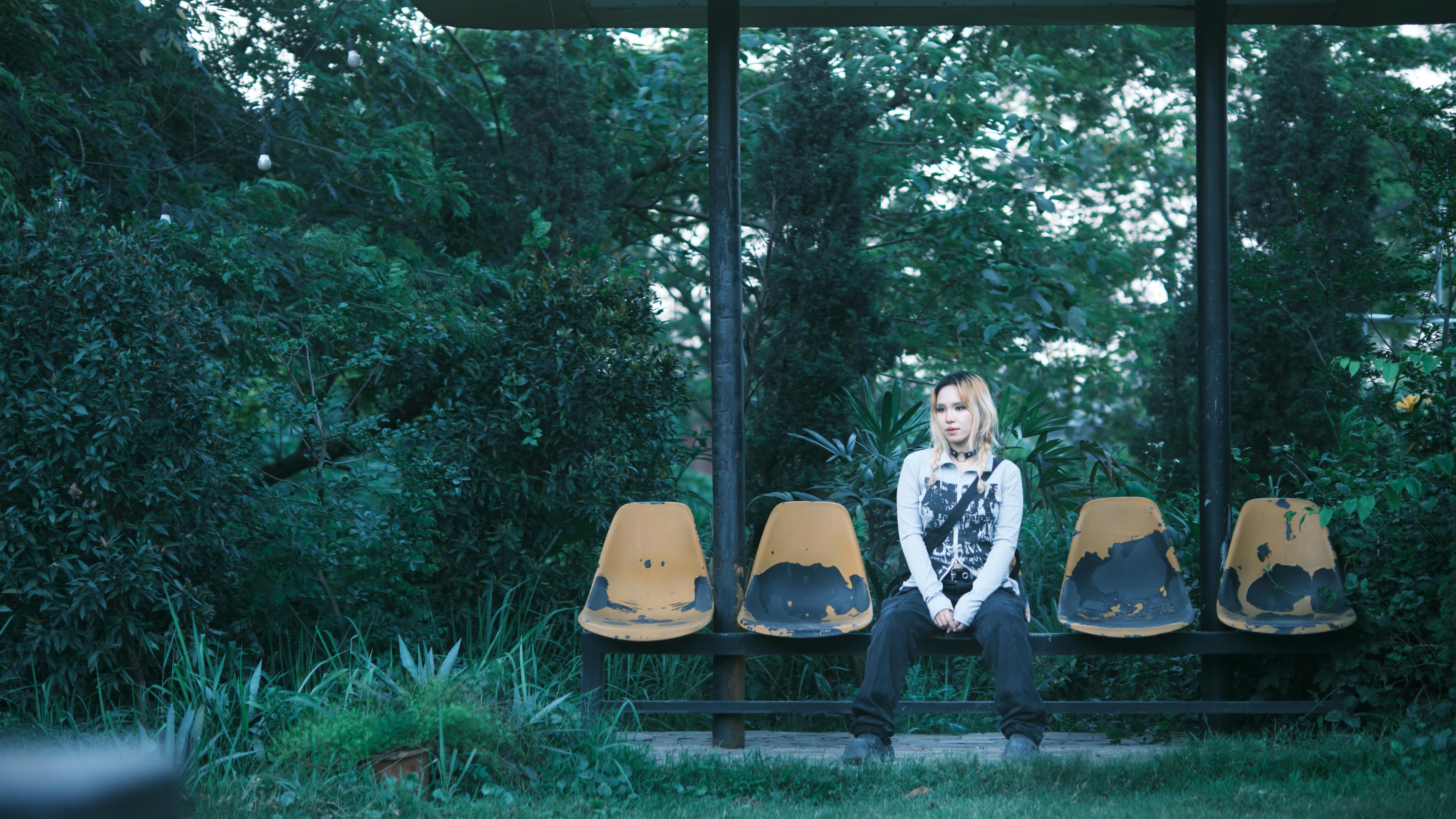 ingyenes Nő egyedül ül egy kopott kültéri padon, buja növényzettel körülvéve. Stockfotó