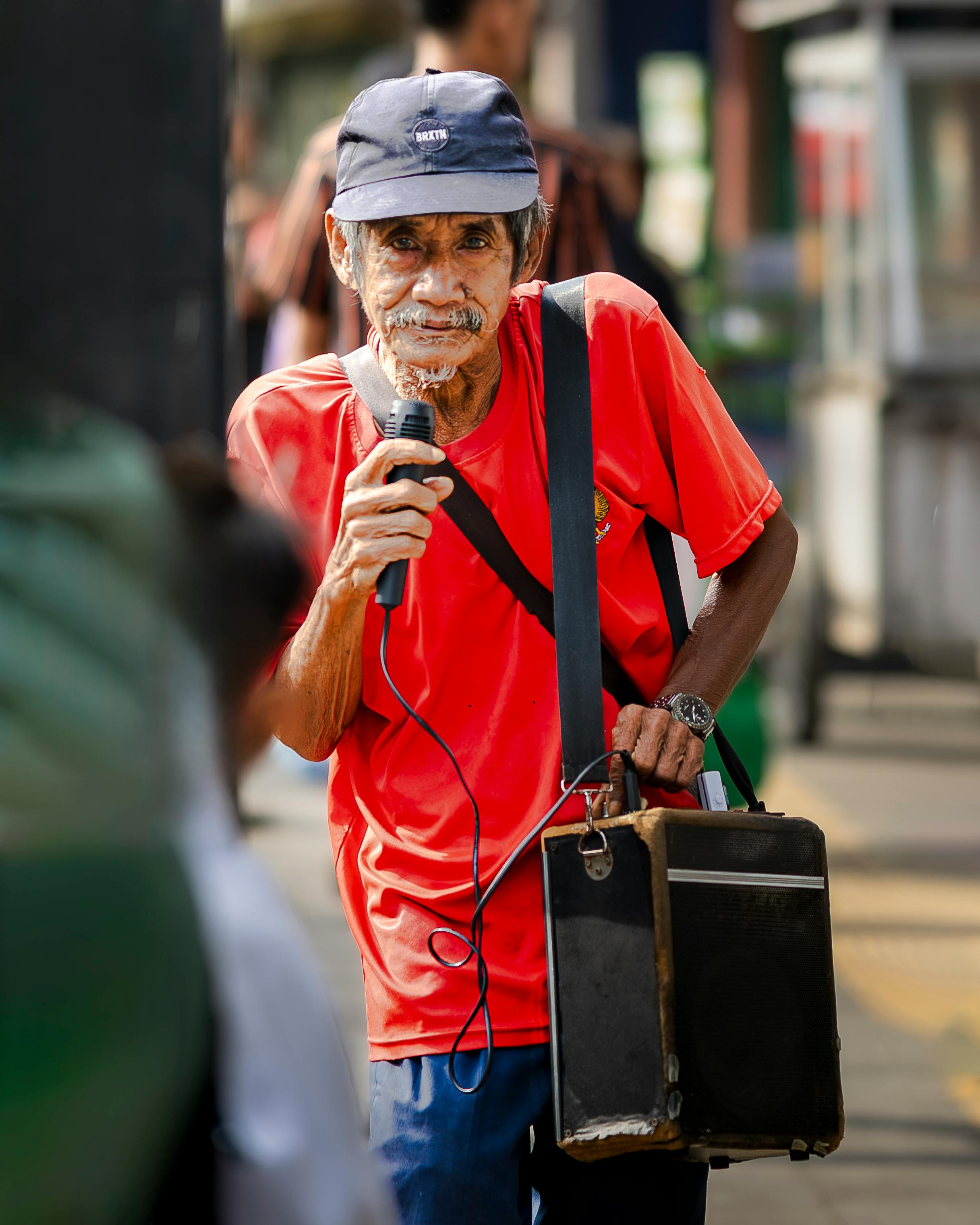 Zadarmo Starší pouličný umelec spievajúci do mikrofónu, oblečený v červenom tričku, vonku v mestskom prostredí. Fotka z fotobanky