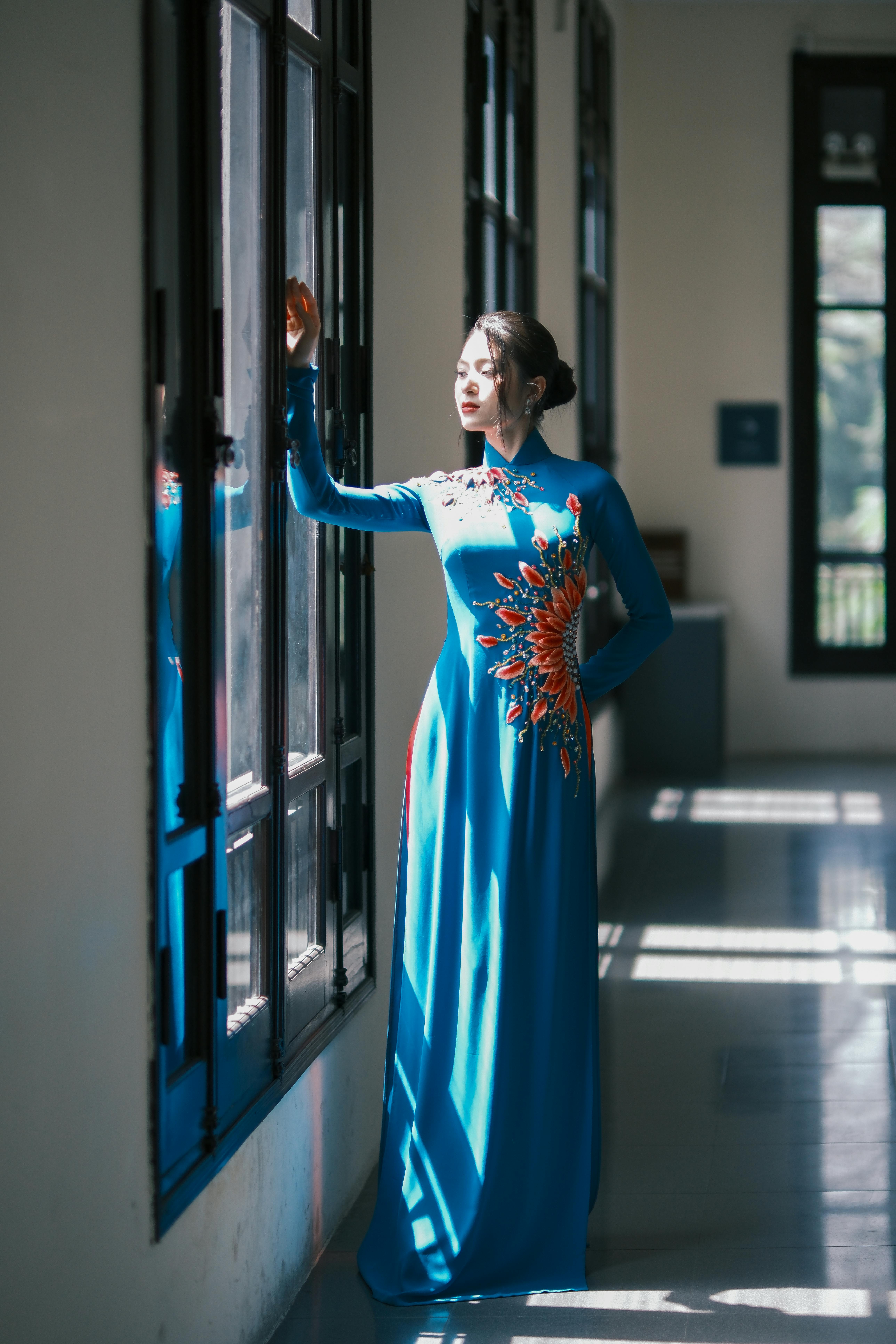 무료 전통적인 파란색 드레스를 입은 여인이 햇살을 받으며 창가에 서 있어 고요한 분위기를 자아낸다. 스톡 사진