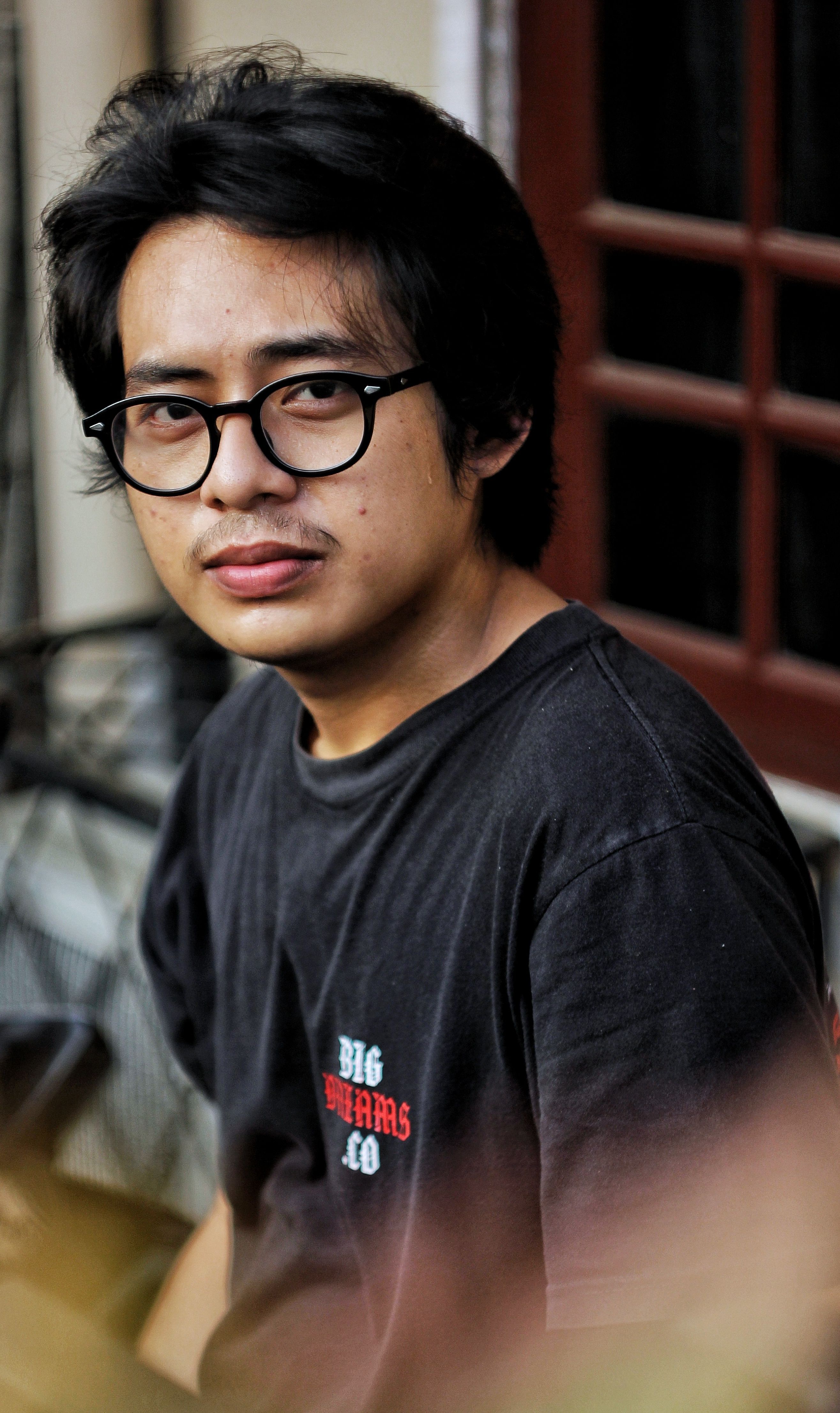 Casual portrait of a young man wearing glasses, looking at the camera in an outdoor setting.