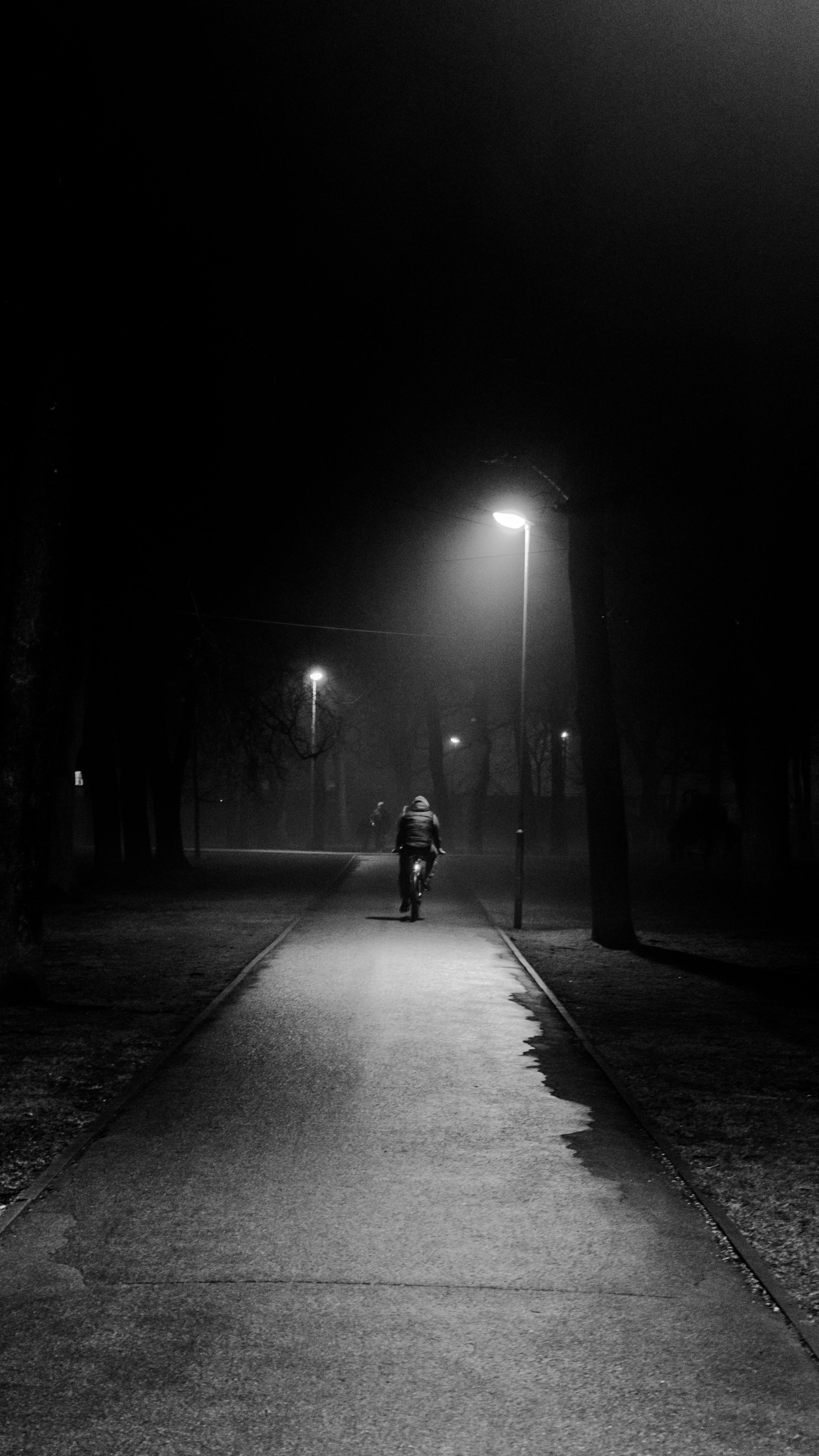 Kostenlos Stimmungsvolles Schwarz-Weiß-Foto, das eine einsame Gestalt zeigt, die nachts unter Straßenlaternen in einem Park in Riga entlanggeht. Stock-Foto