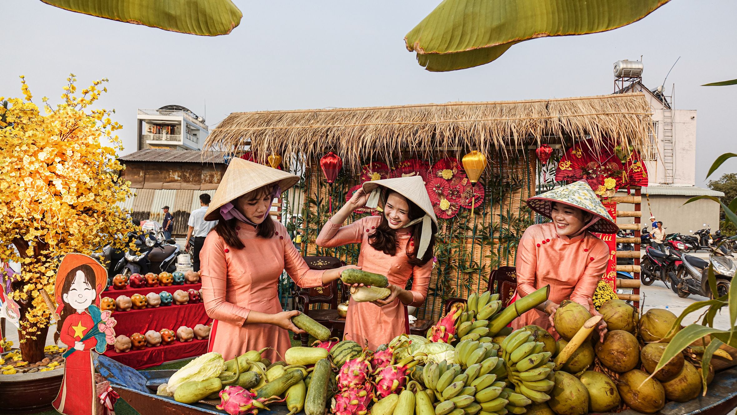 Three women in traditional attire at an outdoor Vietnamese market stall filled with tropical fruits.