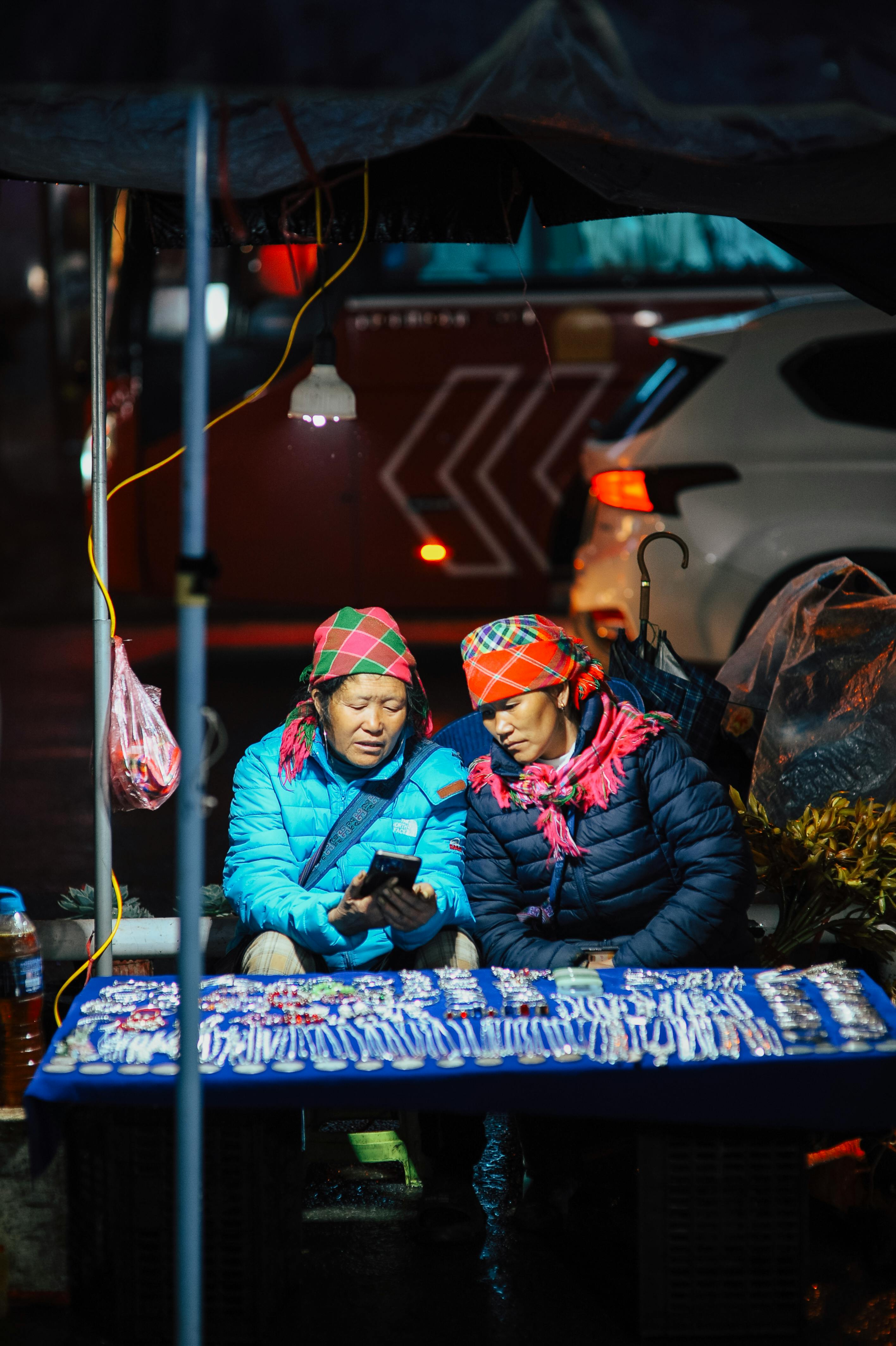 免费 夜市里，两名街头小贩在暖光灯光下用智能手机查看商品。 素材图片