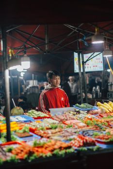 A bustling night market scene featuring a vendor in a red jacket amidst colorful produce.
