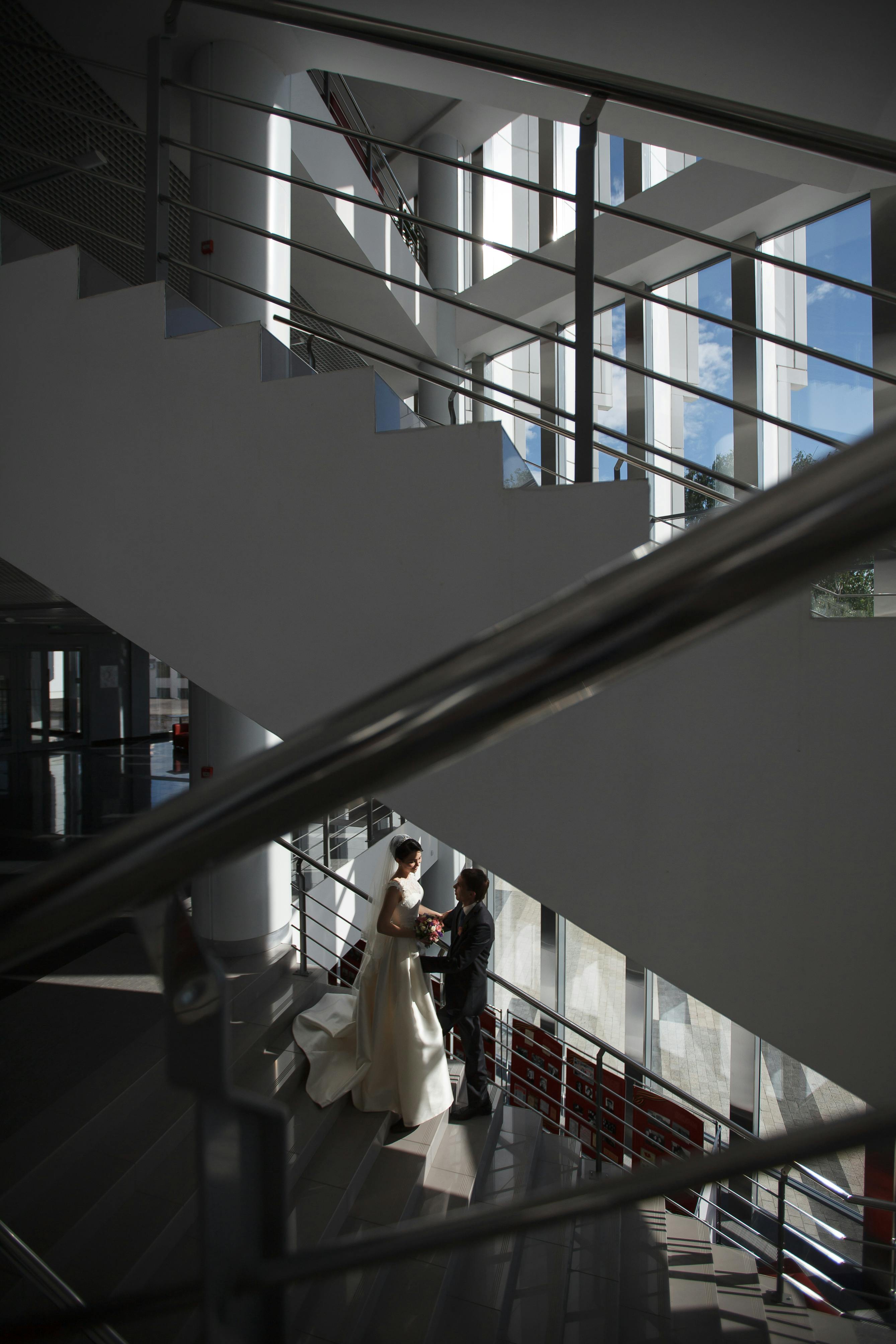 ฟรี เจ้าบ่าวและเจ้าสาวใช้เวลาร่วมกันบนบันไดสไตล์โมเดิร์นภายในบ้านที่ประดับประดาด้วยแสงไฟอย่างสวยงาม คลังภาพถ่าย