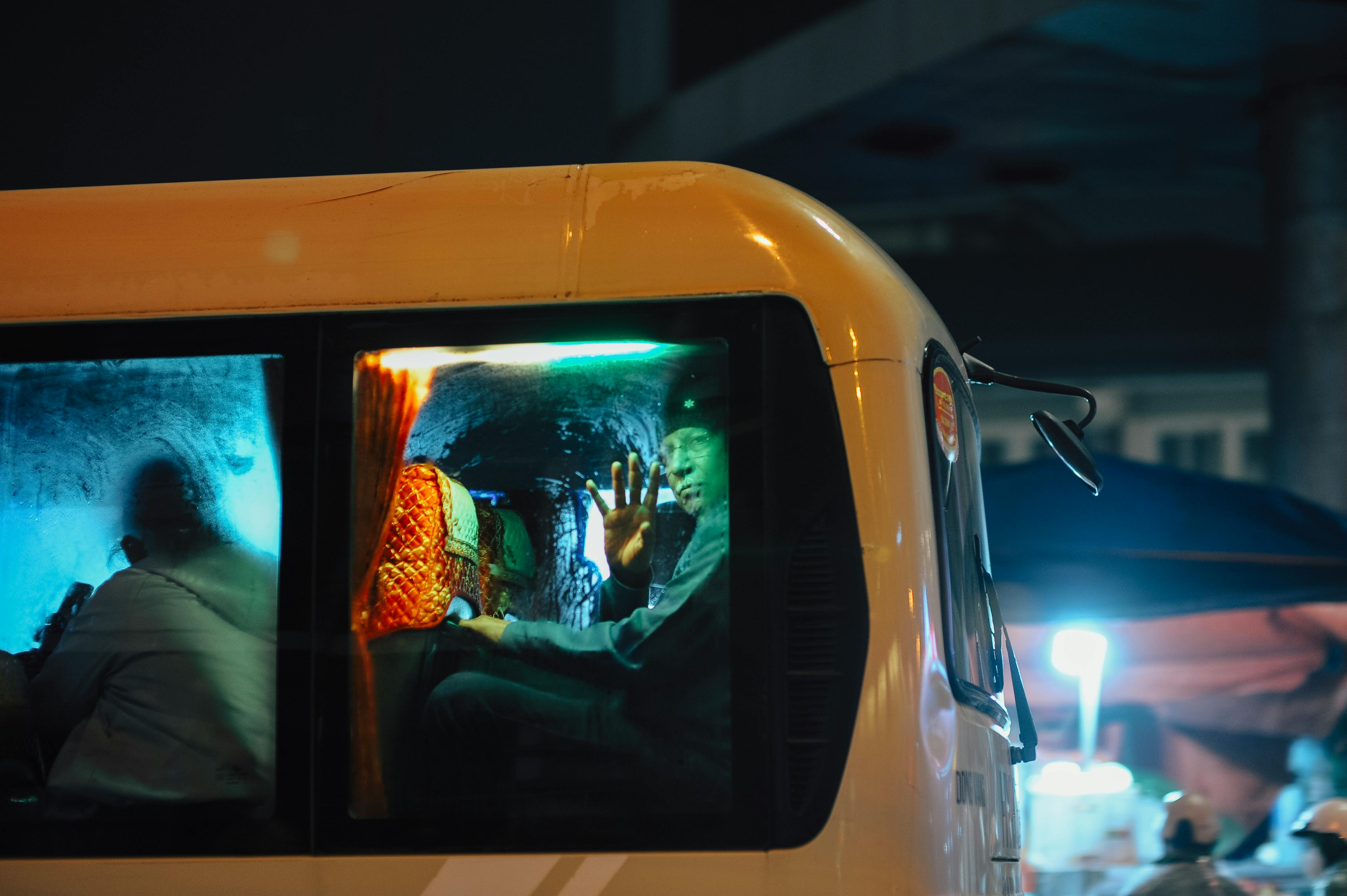 ฟรี คนคนหนึ่งโบกมือจากหน้าต่างรถบัสในเวลากลางคืน โดยมีแสงไฟหลากสีสันส่องสว่างอยู่ภายในรถ คลังภาพถ่าย