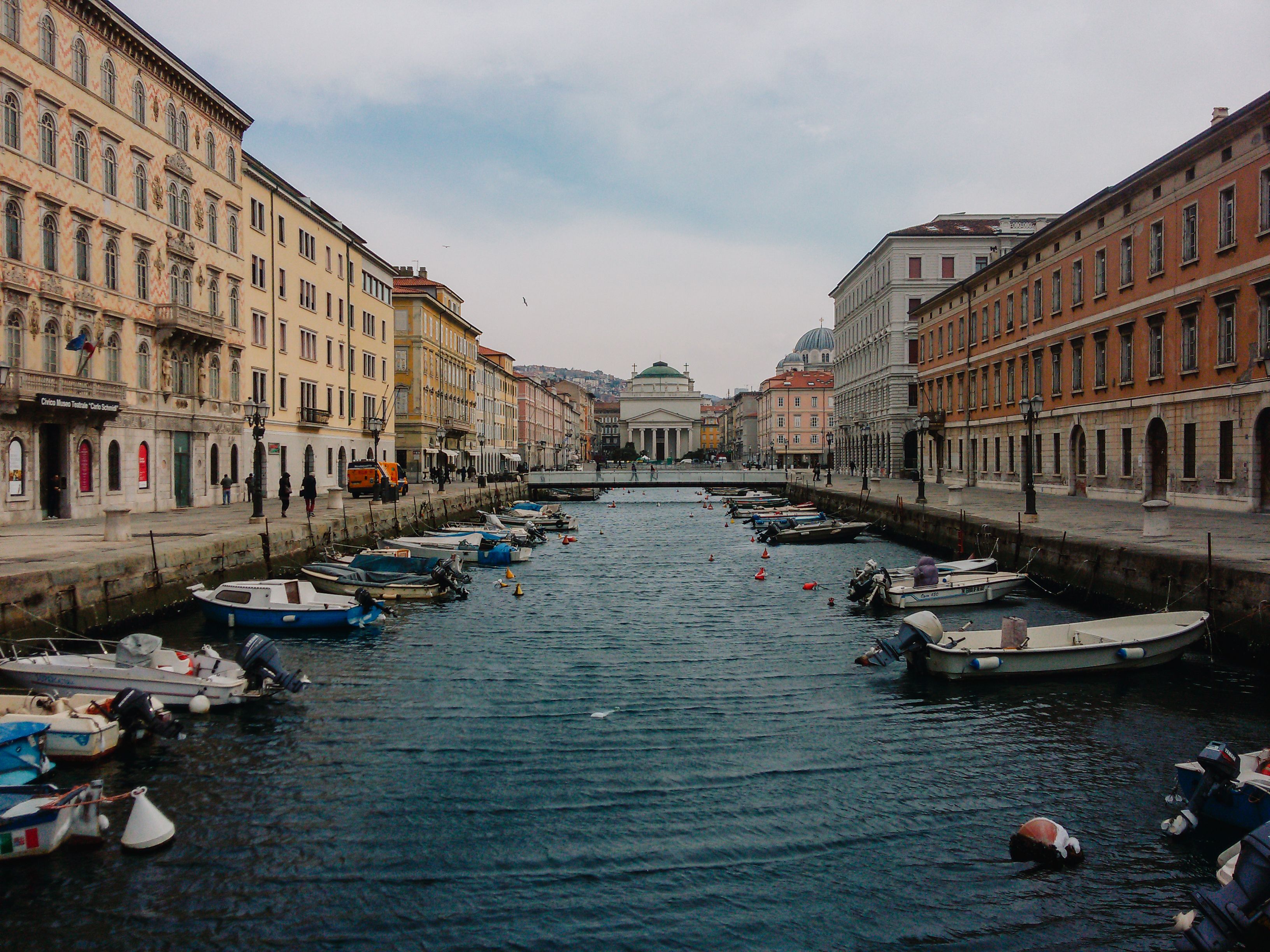 View of Canal Grande and historic buildings in Trieste, Italy with moored boats under an overcast sky.