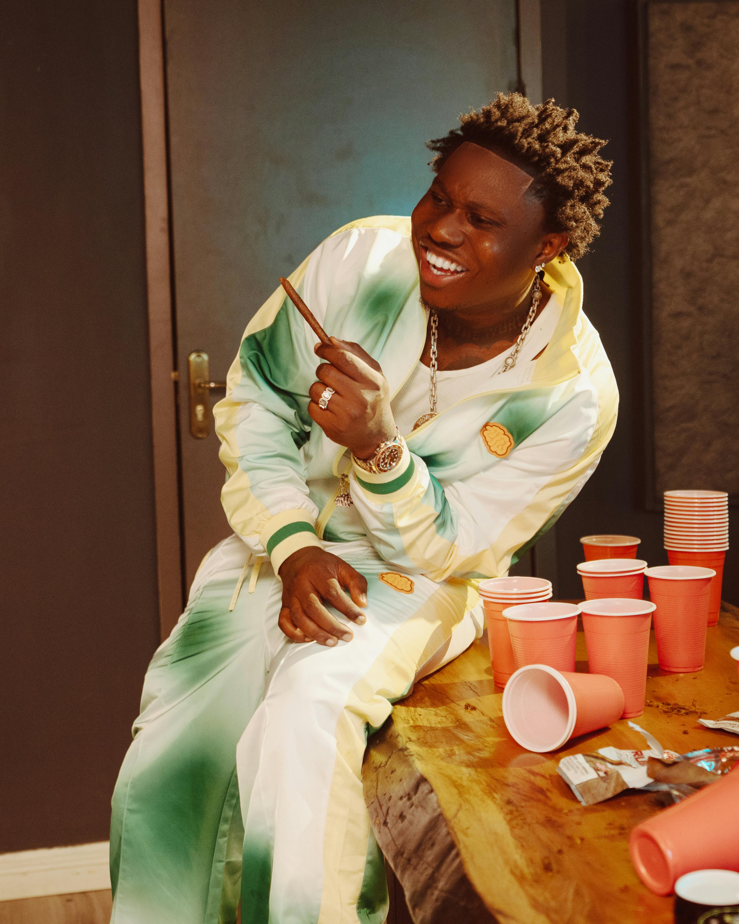 免費 一名身著色彩鮮豔服裝的男子在擺有紅色杯子的宴會桌旁，一邊大笑一邊抽著雪茄。 圖庫相片