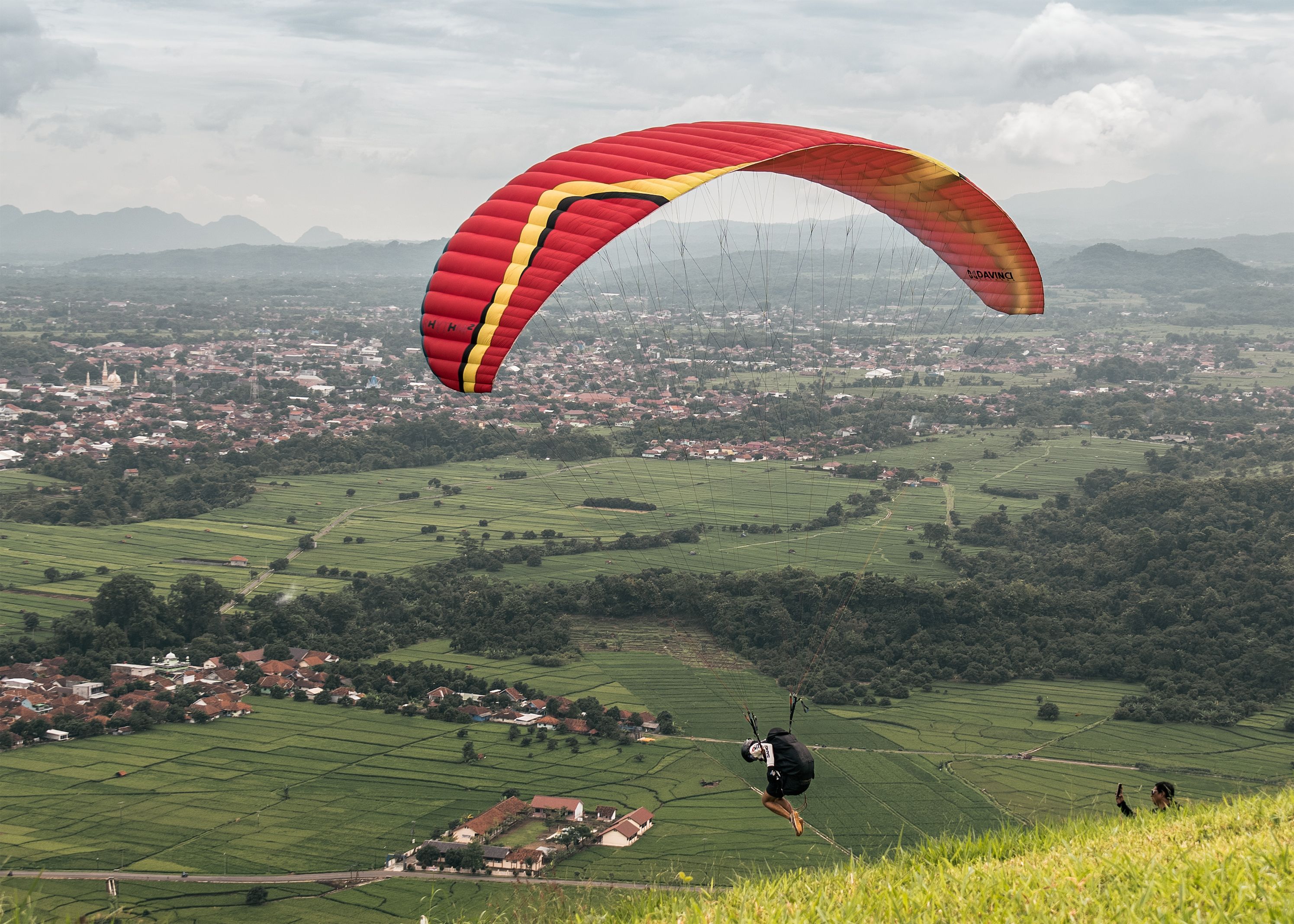 Paraglider soaring over lush landscapes in West Java, Indonesia, with a view of villages and mountains.