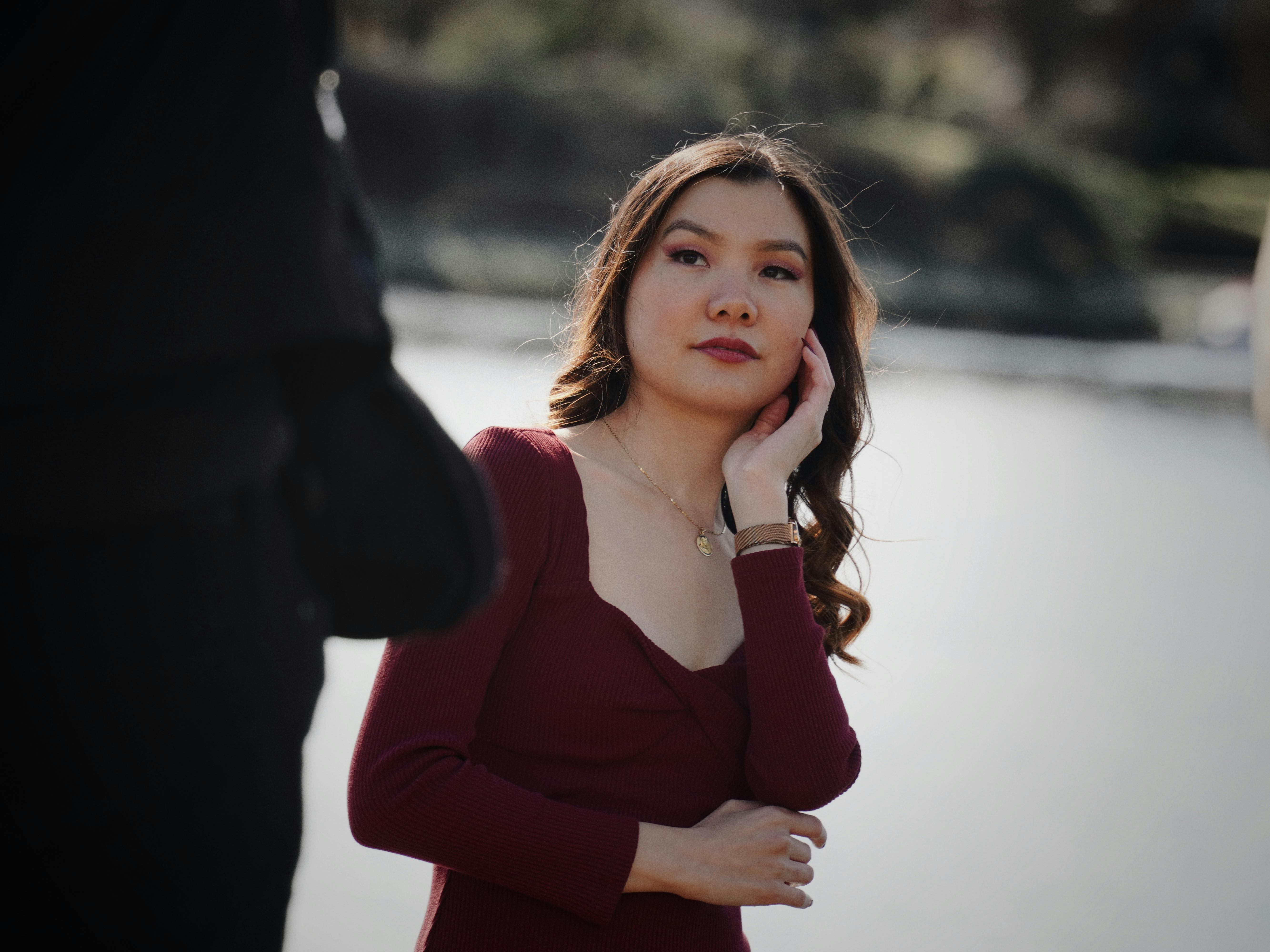 Безкоштовне Молода жінка в бордовій сукні стоїть біля спокійного озера, підкреслюючи свою природну красу. стокове фото