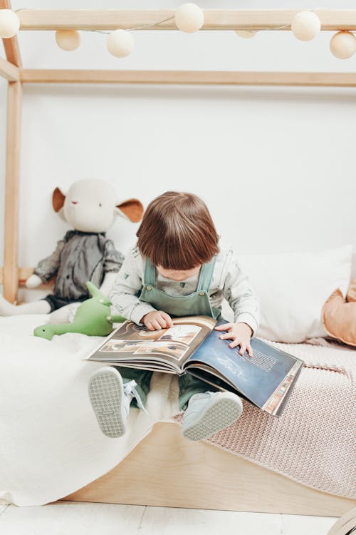 Child in White Long-sleeve Top and Dungaree Trousers Sitting on Bed Reading Book