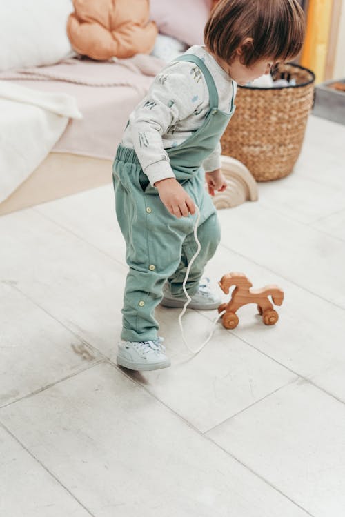 Child in White Long-sleeve Top and Dungaree Trousers Playing With Wooden Toy on White Floor