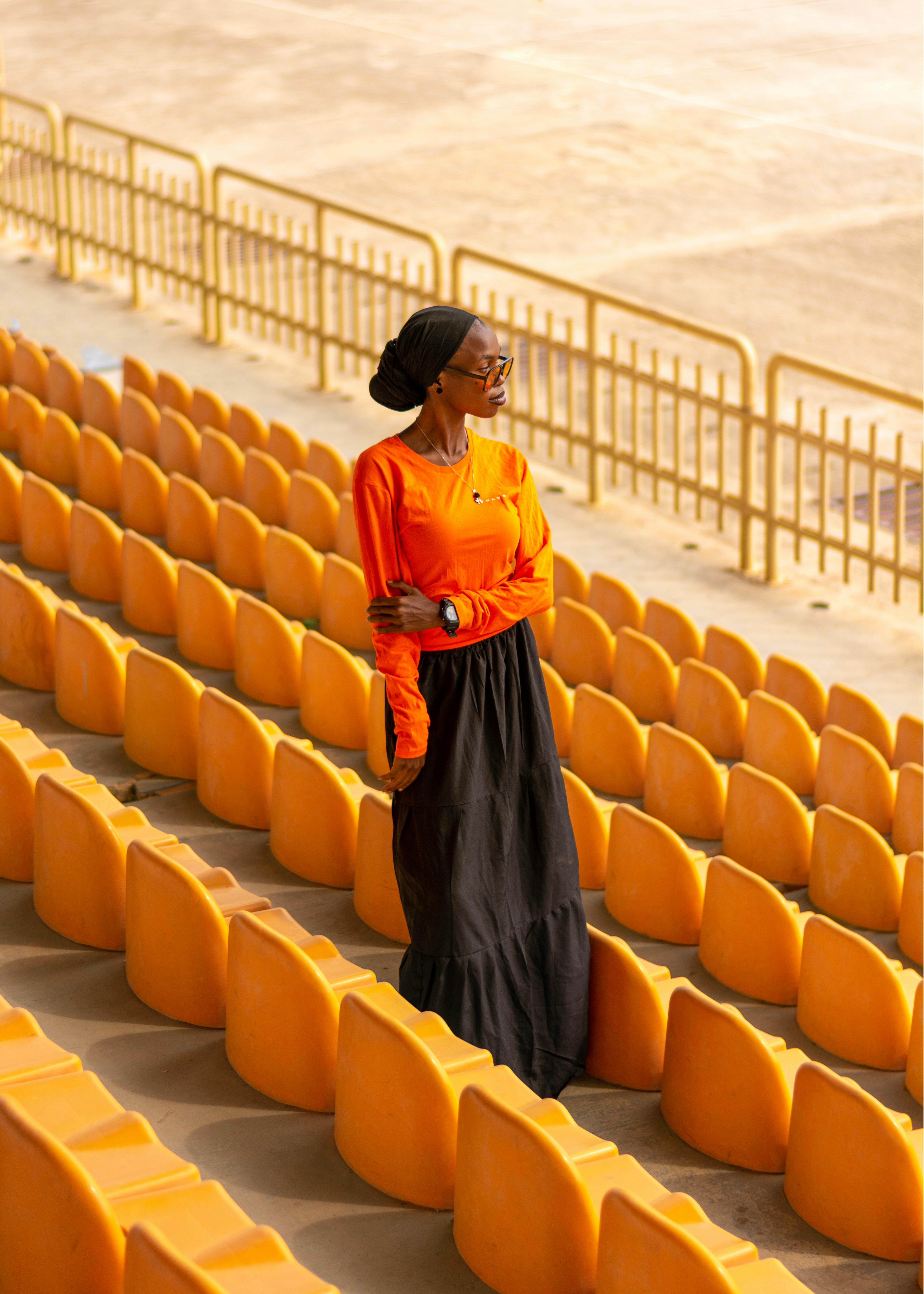 무료 오렌지색 셔츠와 검은색 스커트를 입은 세련된 여성이 텅 빈 경기장 좌석에 서 있다. 스톡 사진