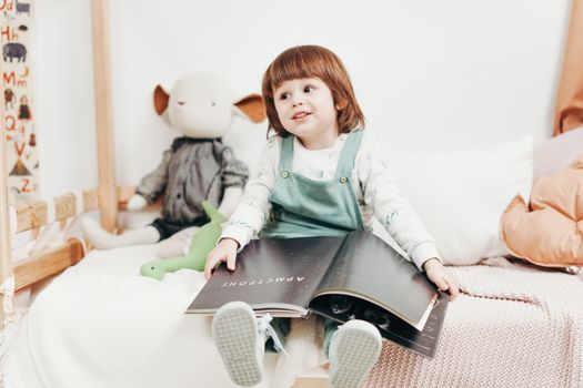 Adorable child sitting on a bed, enjoying a book in a bright playroom with toys.