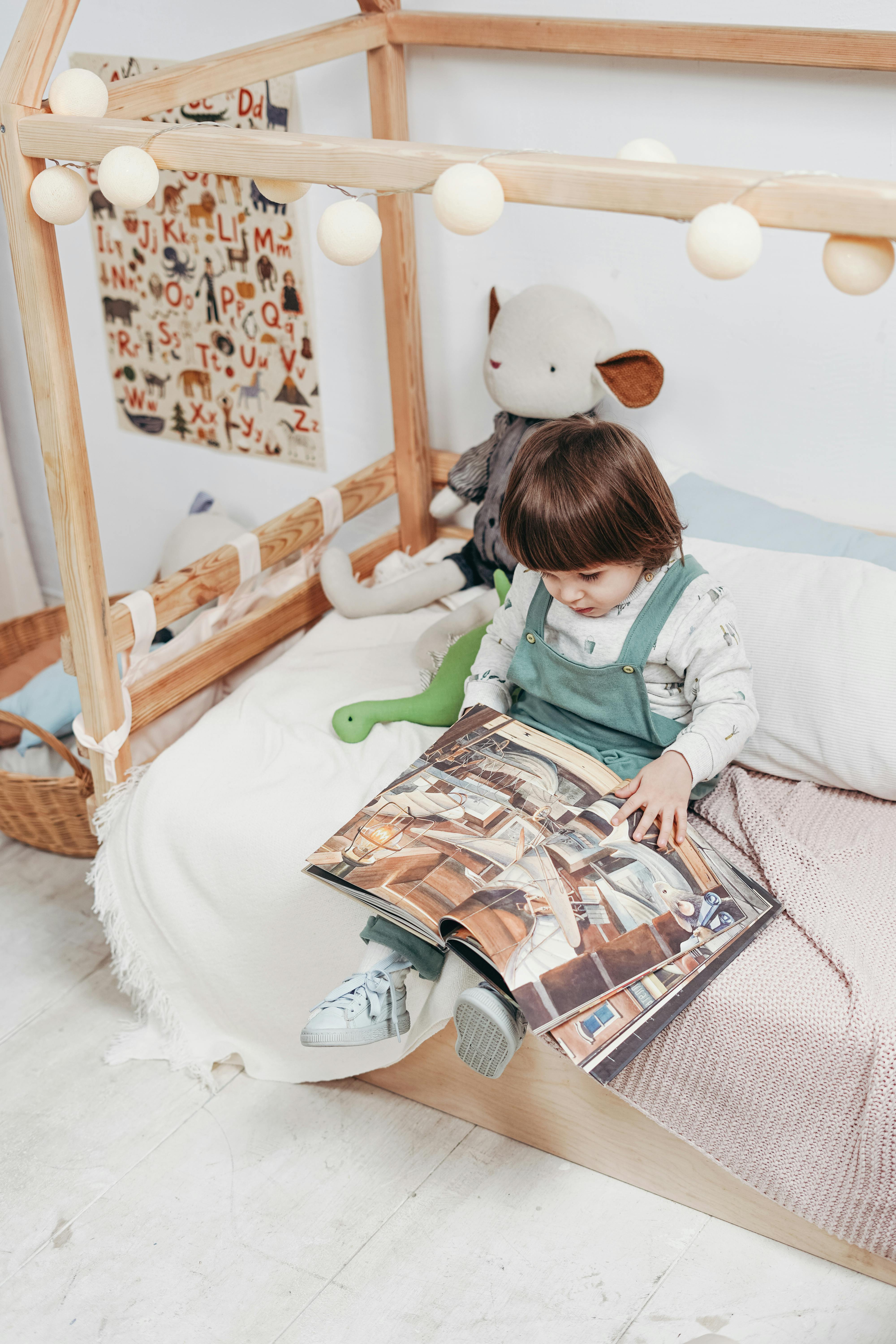 child in white long sleeve top reading book sitting on bed