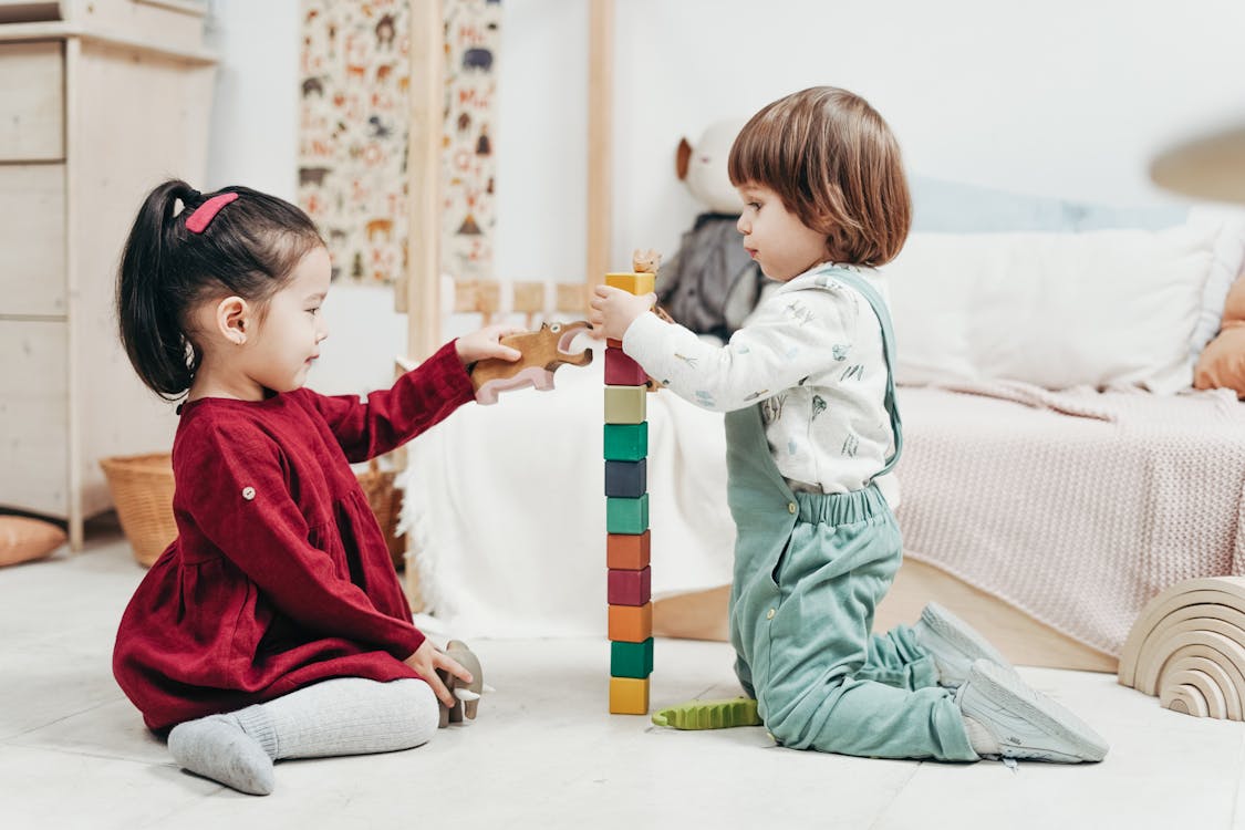  Two children, a girl and a boy, are playing with a variety of colorful wooden blocks and animal figures on the floor in a bedroom.