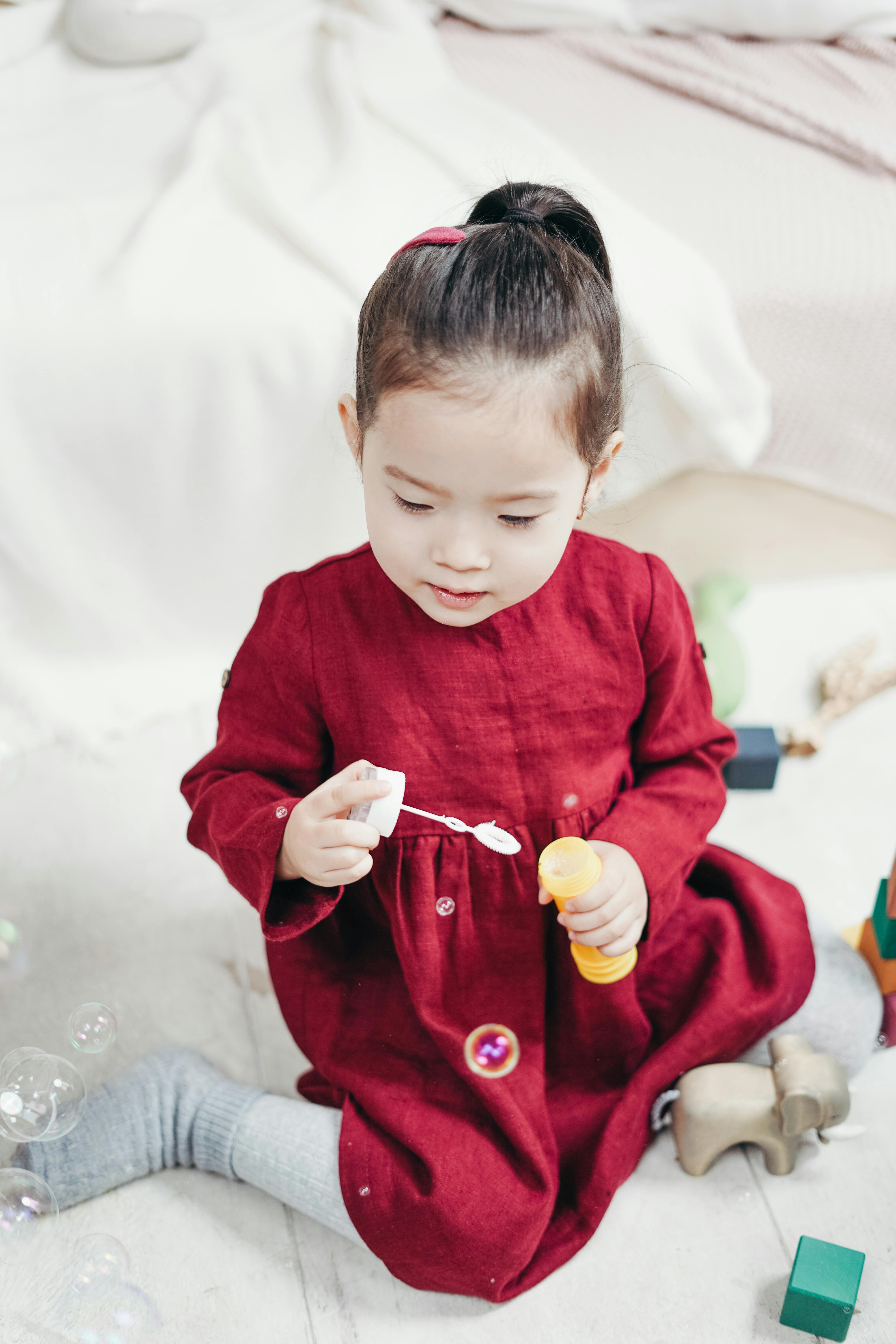 girl in red long sleeve dress sitting on white floor tiles playing with bubble blower